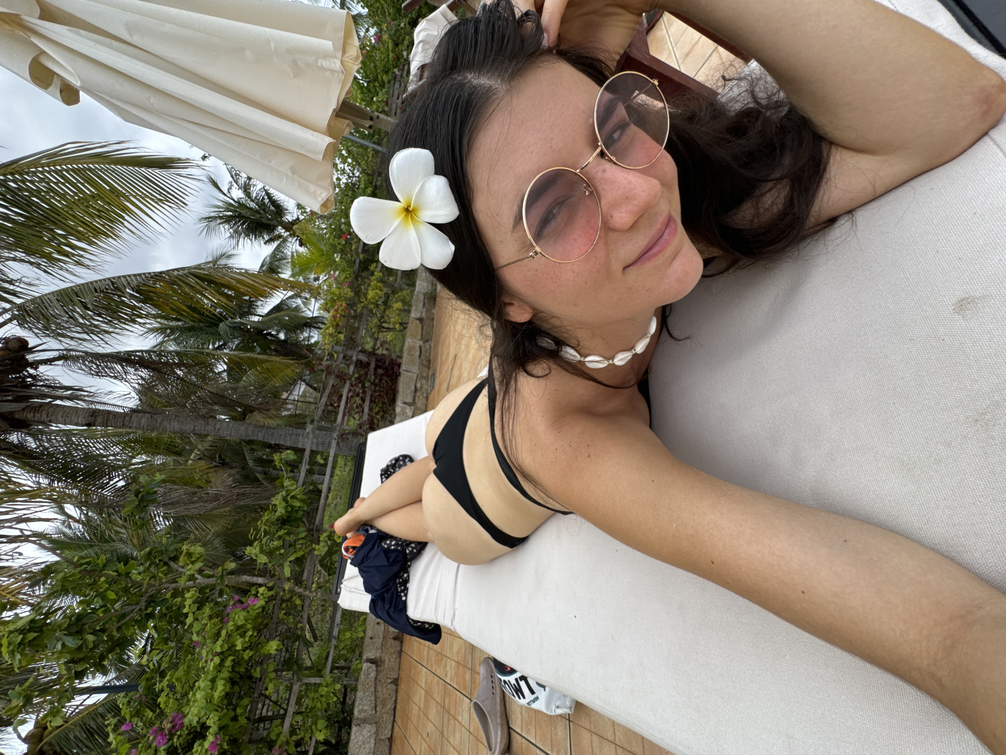 The media file captures a young woman relaxing outdoors on a lounge chair, likely at a tropical resort in Phu Thuy Ward, Vietnam. She is depicted lying on her stomach, facing towards the viewer with a slight smile. She wears a black bikini top and bottoms, round gold-rimmed sunglasses with pink-tinted lenses, and a white shell choker necklace. A white plumeria flower is charmingly tucked behind her left ear. Her dark hair flows around her head.
The woman is engaged in a leisurely activity, possibly sunbathing or simply enjoying the tranquil environment. Her pose and expression suggest contentment and relaxation.
The background is rich with tropical foliage, including tall green palm trees and other leafy plants, some adorned with small pink flowers. The sky above appears overcast or cloudy. In the upper left corner, a large, folded beige parasol is visible. The lounge chair the woman occupies is white and upholstered, and part of a tiled patio or deck is seen to her left. Near her feet on the lounge chair are a dark blue garment, possibly a cover-up, and a gray slip-on shoe. A white bag with the partially visible text "OWT" is also present beside her feet. The overall scene conveys a serene vacation atmosphere in a warm, tropical climate.