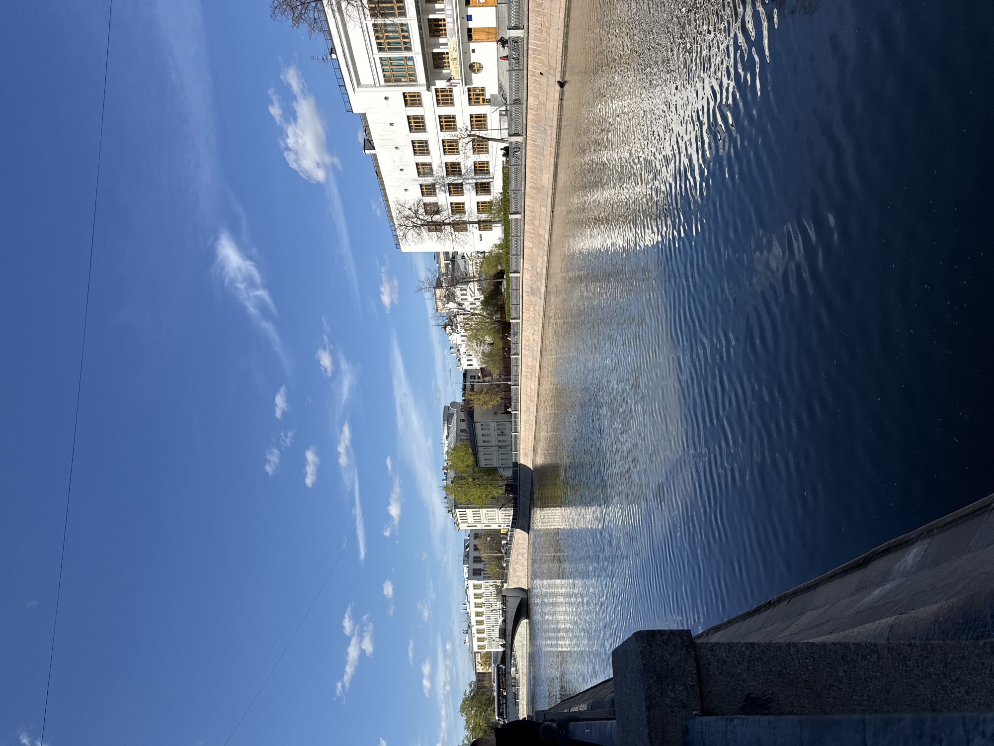 The image depicts a view of the Moskva River in Moscow, Russia, captured from a bridge or embankment. The river reflects sunlight, creating a rippled pattern on the water's surface. Along the riverbank are several white buildings, likely apartments or offices, with visible windows and architectural details. Trees with fresh spring foliage line the embankment, adding a touch of greenery. The sky is blue with scattered white clouds. In the foreground is a stone parapet, suggesting that the viewpoint is from an elevated position such as a bridge or a high embankment.