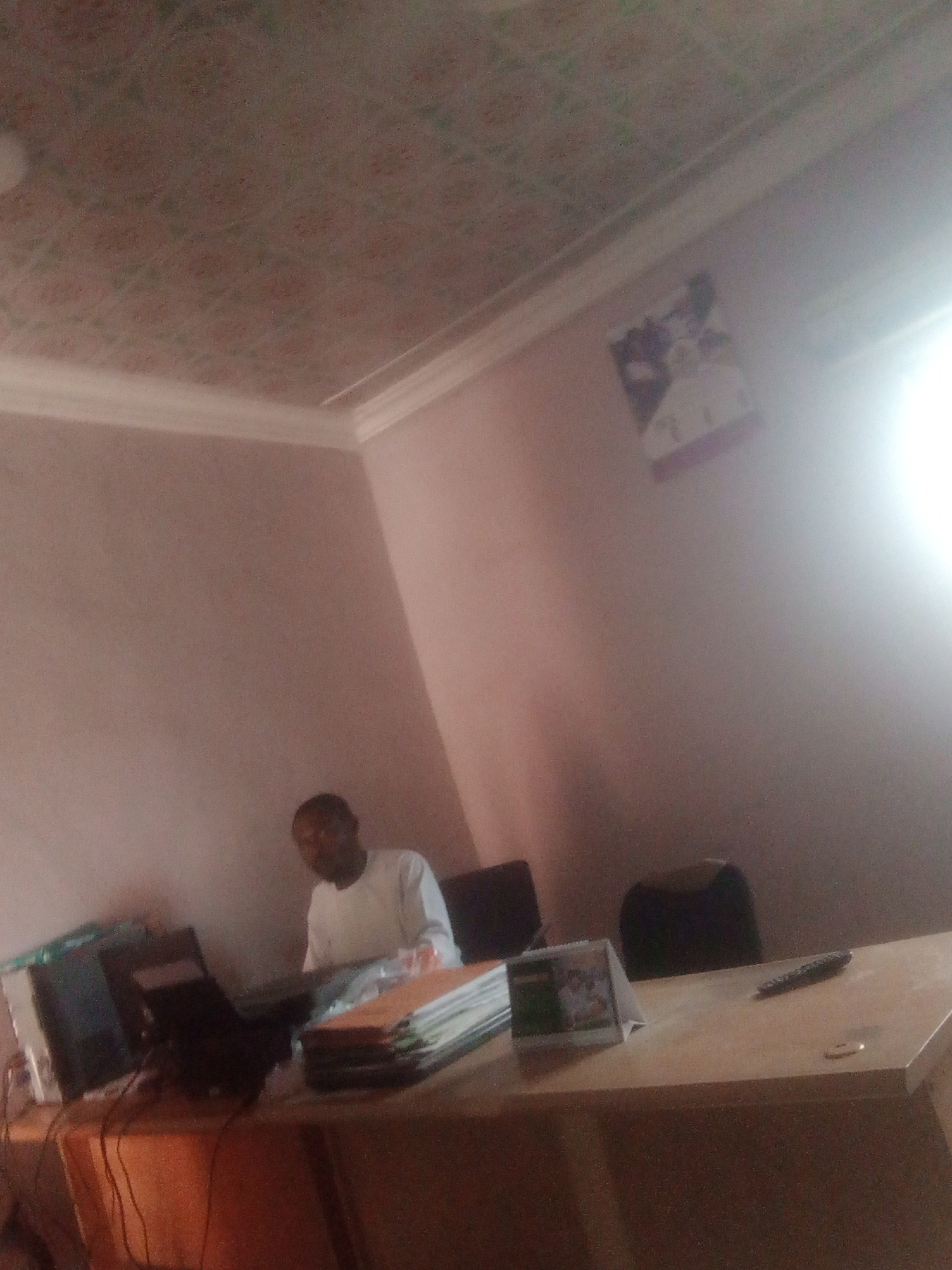 The media file shows an indoor scene, possibly an office, in Jalingo, Nigeria. A man is seated at a wooden desk, wearing a white long-sleeved garment. He appears to be working or interacting with documents on the desk, which are stacked in front of him. To his left, a computer monitor and a laptop are visible, suggesting a workspace.
The room has light pink walls and a patterned ceiling. A framed picture or calendar is mounted on the wall in the background, partially obscured by a bright light source, likely a lamp or natural light entering the room. The lighting is somewhat harsh, creating a glare in the upper right portion of the image.
On the desk, besides the documents and computer equipment, there is a small framed item with what looks like a picture or graphic, and a remote control. A small, circular object, possibly a coin, rests near the remote. The overall impression is that of a functional office environment. The time of day is indeterminate due to the indoor setting and lighting, and no weather cues are present. No visible text is discernable in the image.