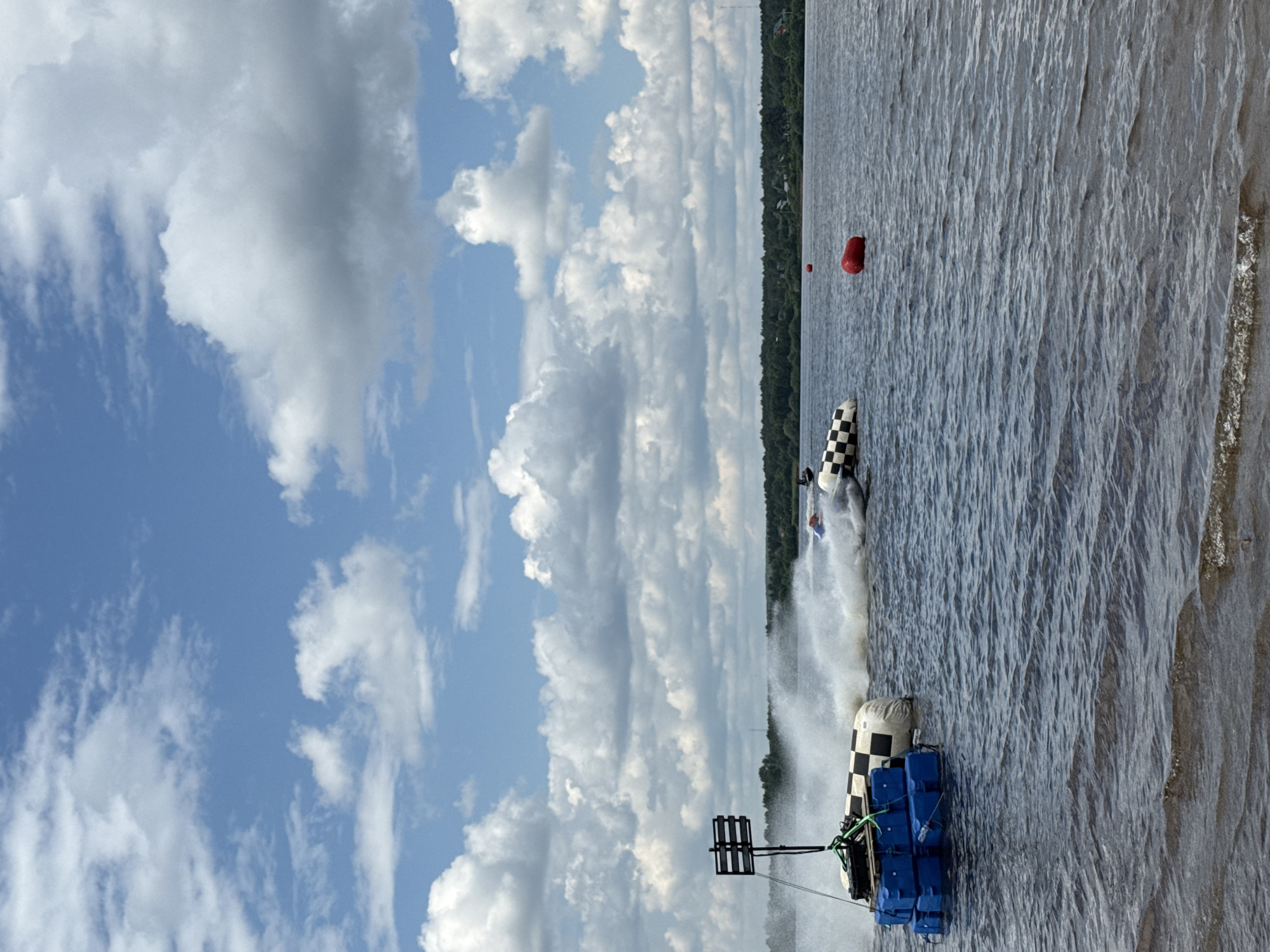 The image depicts a jet ski race on a wide, dark blue body of water, likely a river or lake, under a bright blue sky with scattered white clouds. The foreground shows the dark, rippling surface of the water, with a sandy or muddy shore at the bottom edge. To the left, a black and white checkered buoy is anchored, along with a stack of blue floating containers, and a black marker pole with horizontal bars. In the mid-ground, a jet ski is in motion, creating a large spray of water as it turns, with another jet ski partially visible further to the right. Red buoys are strategically placed in the water, marking a course. In the background, a dense line of dark green trees forms the shoreline, with hints of small buildings or structures nestled within them. The overall scene suggests an outdoor sporting event taking place in a natural setting. Given the context provided, this event is likely occurring on the Kama River in Perm, Russia, which is known for its wide expanse and potential for water sports.