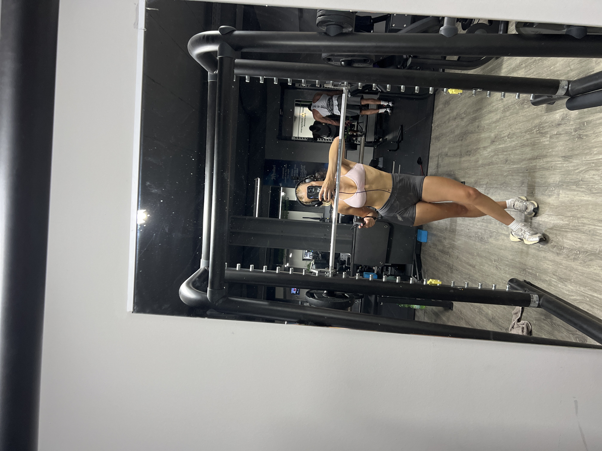 A person is reflected in a large mirror in a gym. The person is wearing a light pink sports bra, grey shorts, white socks, and white athletic shoes. They are holding a smartphone in their left hand and a black object, possibly headphones, in their right hand, and taking a selfie. The person is leaning against a black Smith machine. In the background, other gym equipment and two other people can be seen. The floor is made of light wood-colored laminate. The lighting is artificial. The city of Altötting, Germany, is not discernible from the image.