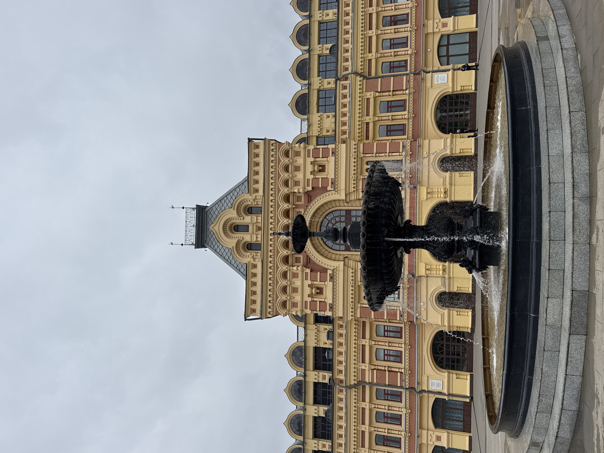 A large, ornate black fountain with water spraying upwards and outwards is in the foreground of the image. The fountain is situated in a paved plaza in front of a grand, yellow and red building. The building features multiple arched windows and decorative architectural elements, with a grey, mansard-style roof and a central tower. Two individuals are visible in the background near the building's entrance. The sky above is overcast and grey. This is likely the Nizhny Novgorod State Academic Opera and Ballet Theatre named after A.S. Pushkin, located in Nizhny Novgorod, Russia.