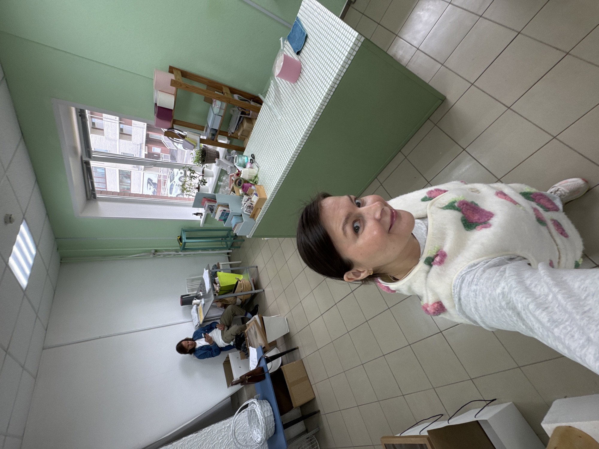 An indoor scene in Izhevsk, Russia, is captured from a selfie perspective. The main subject is a woman in the foreground, taking the photo. She has dark hair and a neutral expression, looking towards the camera. She is wearing a white, fluffy vest adorned with pink and green floral designs, over a grey long-sleeved shirt.
The setting appears to be a multi-purpose room, possibly a workshop, small office, or craft store. A long, light green counter with a white grid-patterned top dominates the midground. Various items are arranged on the counter, including a pink cylindrical container, a blue cloth, small potted plants, decorative objects, and what seems to be stationery or craft supplies displayed on shelves. Behind the counter, a wooden shelving unit holds more items.
In the background, another person, possibly a man, is seated at a grey metal desk. He wears a blue denim jacket over a white t-shirt and olive green pants, and appears to be occupied with a phone or object in his hands. His desk is equipped with a computer monitor, papers, and a yellow bag. Cardboard boxes and a white wicker basket are visible on the tiled floor near his workspace.
The room features light green walls, a white ceiling with recessed light panels, and light-colored tiled flooring. A large window on the left side of the frame allows ample natural light into the space, suggesting it is daytime. Through the window, parts of urban buildings with visible windows can be seen, indicating an outdoor city environment. No discernible text is visible in the image. The overall atmosphere suggests a casual, possibly work-related or hobby-oriented environment.