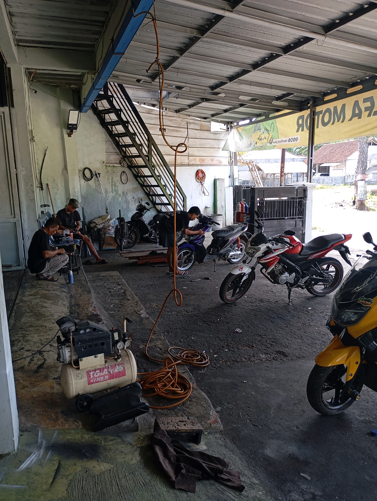The image depicts an open-air motorcycle repair shop in Sodonghilir, Indonesia. Three men are actively engaged in motorcycle maintenance. One man, wearing a grey cap and dark t-shirt, is seated on a stool, working on a motorcycle. Another man, in a dark t-shirt and black pants, is kneeling on the ground, also attending to a motorcycle. A third man, dressed in a black t-shirt and shorts, is leaning over a motorcycle in the background. Several motorcycles are present, including a red and white sportbike, a dark colored motorcycle, and the front portion of a yellow motorcycle is visible in the foreground. An air compressor with the label "TGA 125" is situated on the ground, along with coiled orange hoses and other tools. The workshop floor appears to be concrete and is somewhat dusty. Overhead, a metal roof structure supported by beams is visible, along with a staircase leading to an upper level. A banner with text is hanging on the wall. The overall impression is of a busy and functional repair shop.
