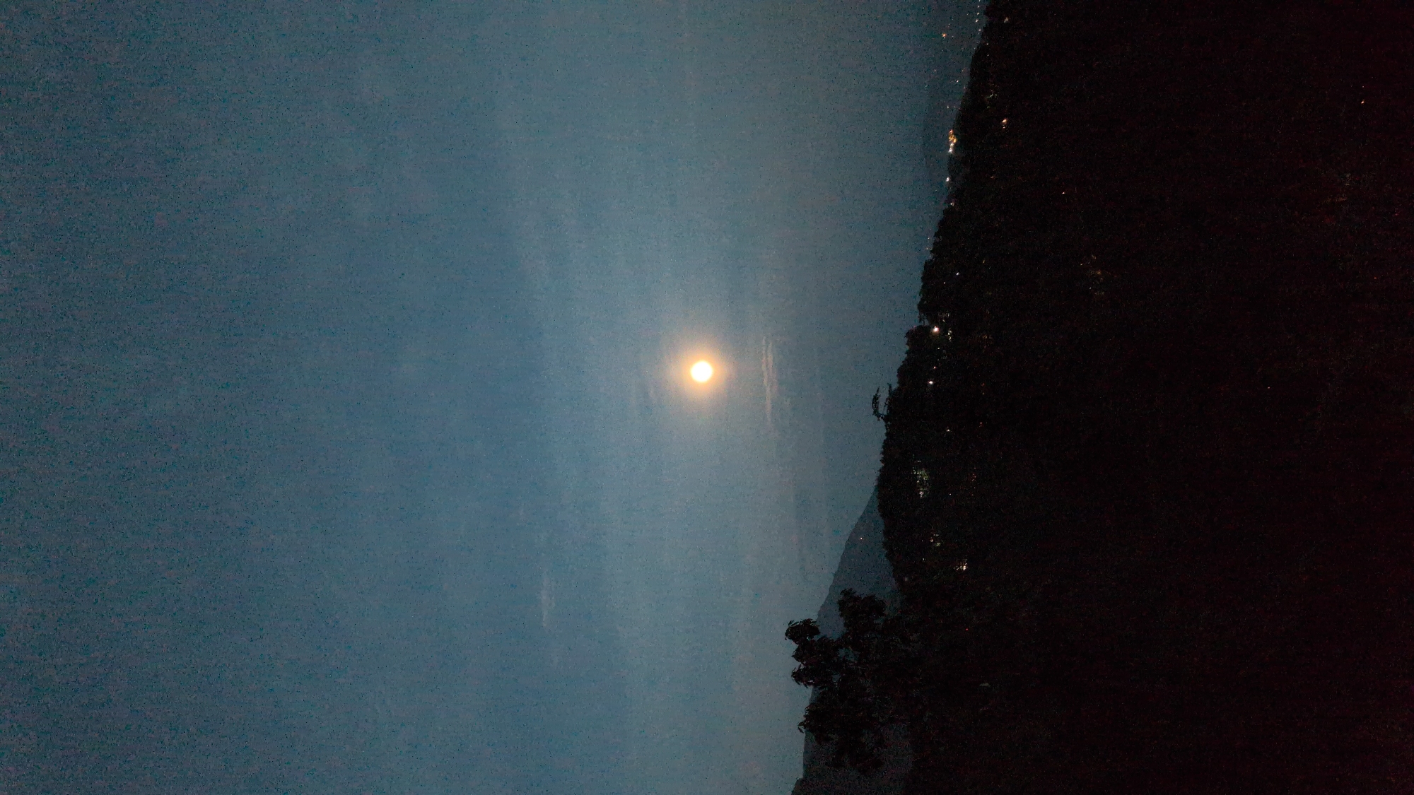 The image depicts a nighttime scene, possibly a landscape, in Dharamkot, India. The sky is a dark, muted blue, with a distinct, bright circular light source, likely the moon. The terrain on the right is dark and densely shadowed, suggestive of a hillside with trees or dense vegetation. Small points of light are scattered along the upper portion of the hillside, hinting at the presence of buildings or settlements. The overall atmosphere is quiet and subdued, with a sense of remoteness due to the limited visibility and lighting conditions.