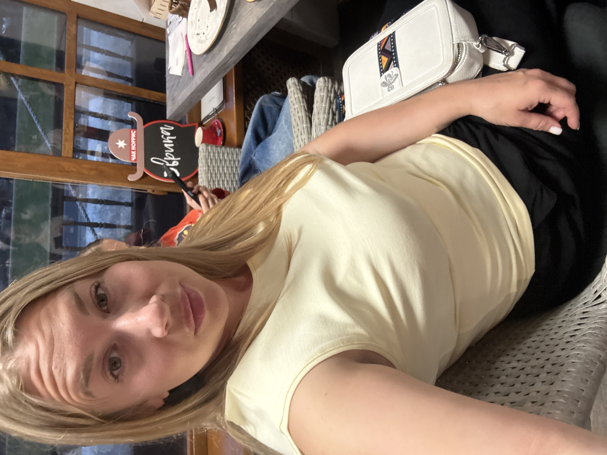 A woman with blonde hair is taking a selfie indoors. She is wearing a light yellow sleeveless top and black pants. A white handbag with a decorative strap is resting on her lap. In the background, a table with a plate, a cup, and a menu are visible. A wooden window with multiple panes is to the right of the woman. The scene appears to be set in a cafe or restaurant.