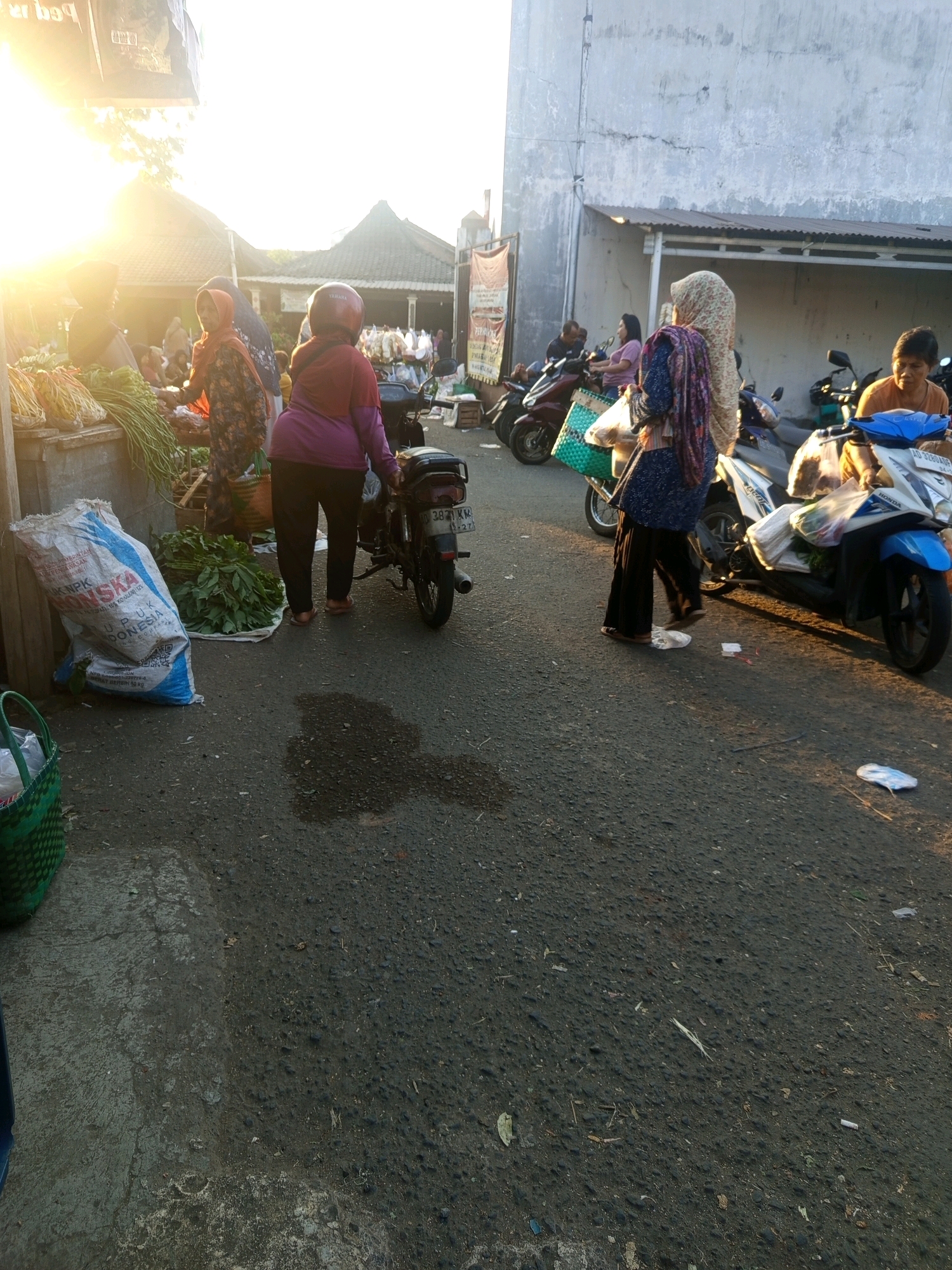 The image depicts a street-level view of a market scene in Tuban, Indonesia, likely during early morning or late afternoon given the low angle of the sun and the warm, diffused light. The scene is populated by several individuals, primarily women, engaged in market activities.
In the foreground, a woman wearing a purple top and black pants, with a red helmet, is standing next to a motorcycle. Her posture suggests she might be about to mount or dismount the vehicle. Nearby, a large white and blue sack, possibly containing fertilizer (indicated by text like "NPK" and "INDONESIA"), is leaning against a low wall or stall. To the left of the sack, a green woven basket is visible.
Further into the scene, other individuals are seen interacting. One woman, wearing a colorful patterned hijab and dress, is carrying a basket and appears to be walking. Another woman, dressed in a light-colored hijab and a dark top, is also present. A few motorcycles are parked along the side of the street, some with items placed on them, possibly for transport or display. The background shows a building with a corrugated metal roof and a light-colored wall, typical of commercial structures in the area. The ground is paved with asphalt and shows signs of being wet in some areas, possibly from recent rain or cleaning. The overall atmosphere is that of a bustling local marketplace.