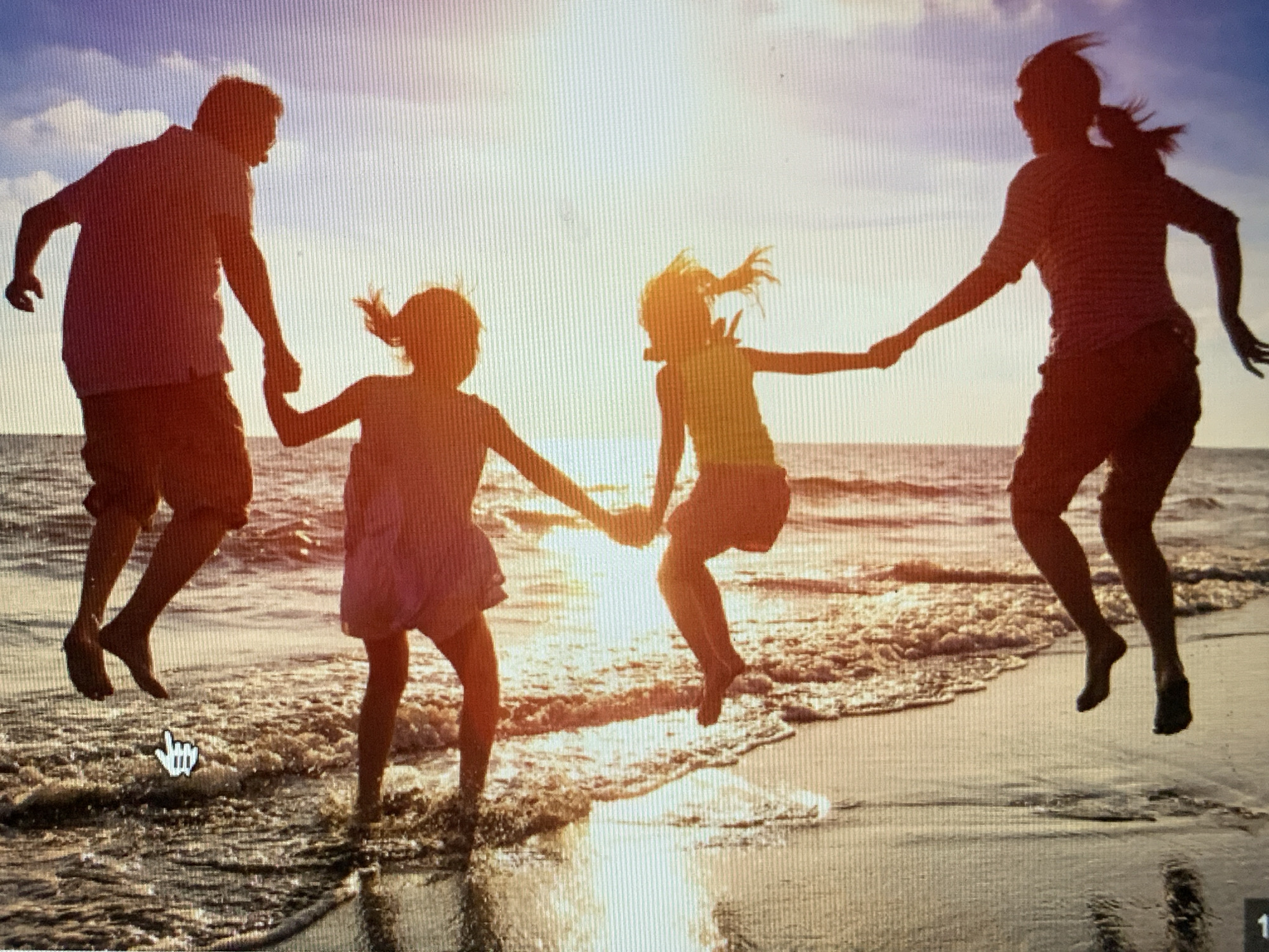 The image shows a family jumping at the beach during sunset. There are two adults, a man and a woman, and two children, likely girls. All four are silhouetted against the bright sun and sky. They are holding hands and jumping in unison, with the water of the ocean reaching their feet. The location is likely a beach but it does not appear to be in Barcelona, Spain. There are no landmarks to suggest the location is Spain.