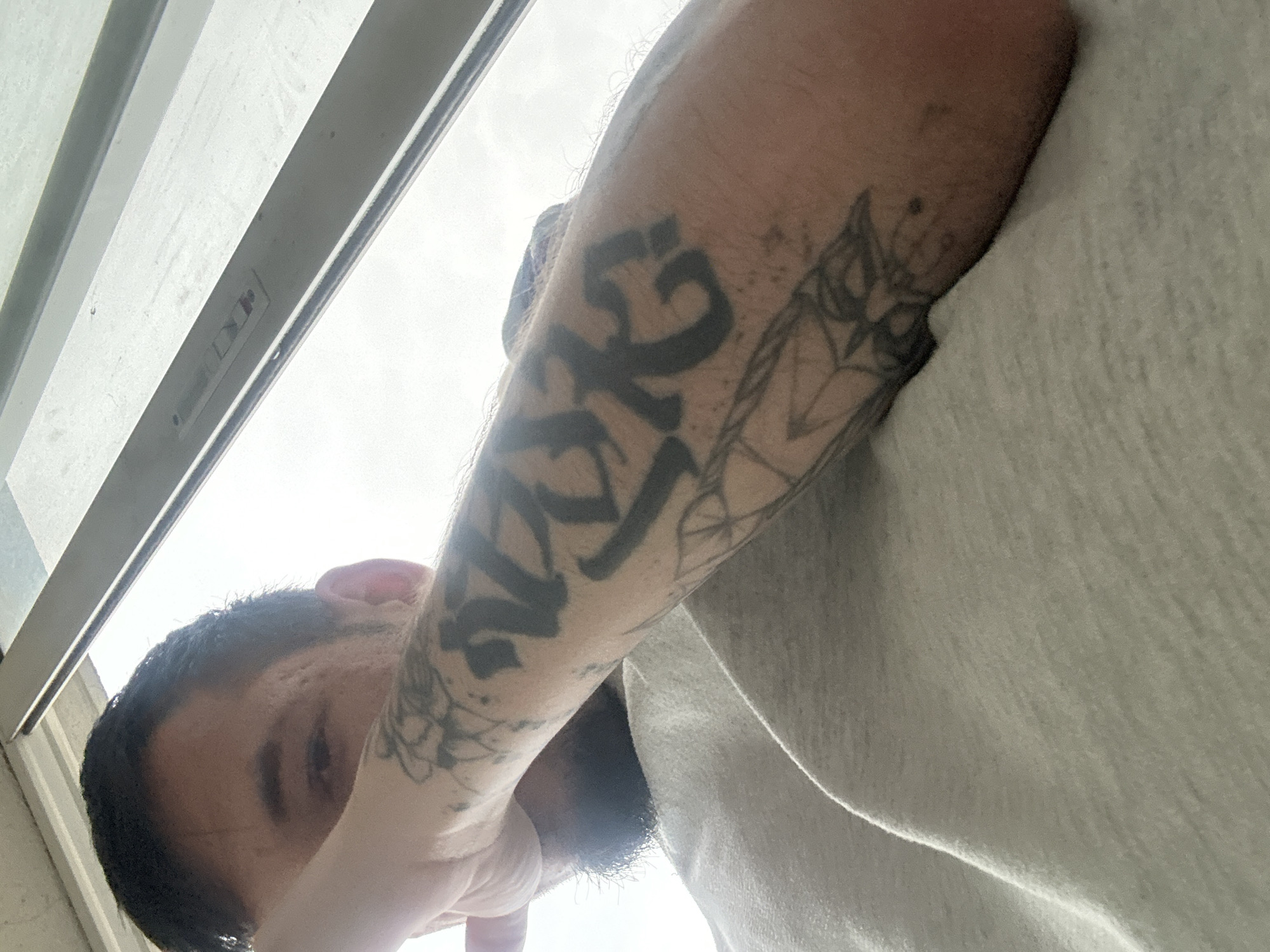 The media file captures a low-angle, upward shot of a person looking towards a brightly lit window. The main subject is a male, identified by his short, dark hair and visible facial hair (beard/stubble). His face is partially obscured by his right arm, which is heavily tattooed.
On his right forearm, a prominent, dark, stylized tattoo spells out what appears to be "NATE" or "NÄTE" in a bold, blocky font. Adjacent to this, further up the arm, are lighter, outline-style tattoos, possibly depicting abstract, floral, or geometric patterns. The skin around the tattoos has visible texture and some fine hair.
The setting appears to be indoors, looking out through a window. A light-colored window frame or sill is visible along the top and left edges of the frame. Bright, diffused light streams in from outside, suggesting an overcast daytime sky. On the right side of the image, a light grey or off-white garment, possibly a sweatshirt or blanket, is visible, adding to the impression of an indoor environment. The person's activity seems to be looking up or observing something through the window from a low vantage point. No specific emotions are discernible due to the obscured face. The location context provided is Varna, Bulgaria.