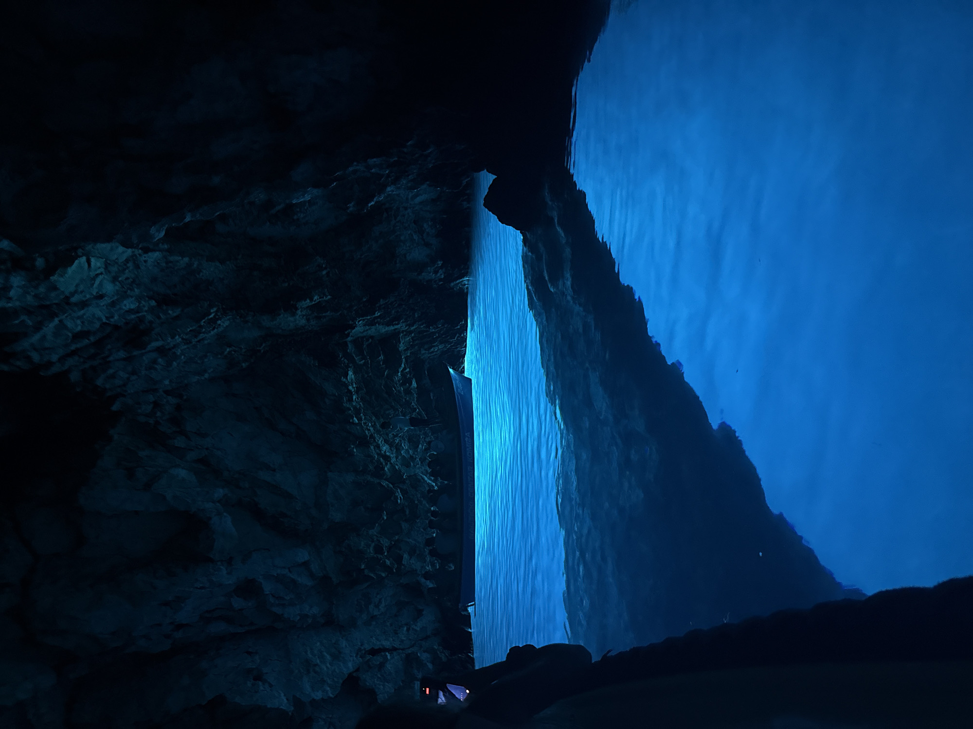 A boat floats on bright blue, illuminated water within a dark cave. The cave walls are rough and rocky, with a dark, shadowy texture. The water reflects the rocky ceiling. A person is visible on the left side of the frame, holding a phone and appearing to take a photo. The boat has people seated inside. The scene is illuminated by an unseen light source that causes the water to glow intensely blue. The location is the Blue Cave on the island of Biševo, near the town of Komiža, Croatia.