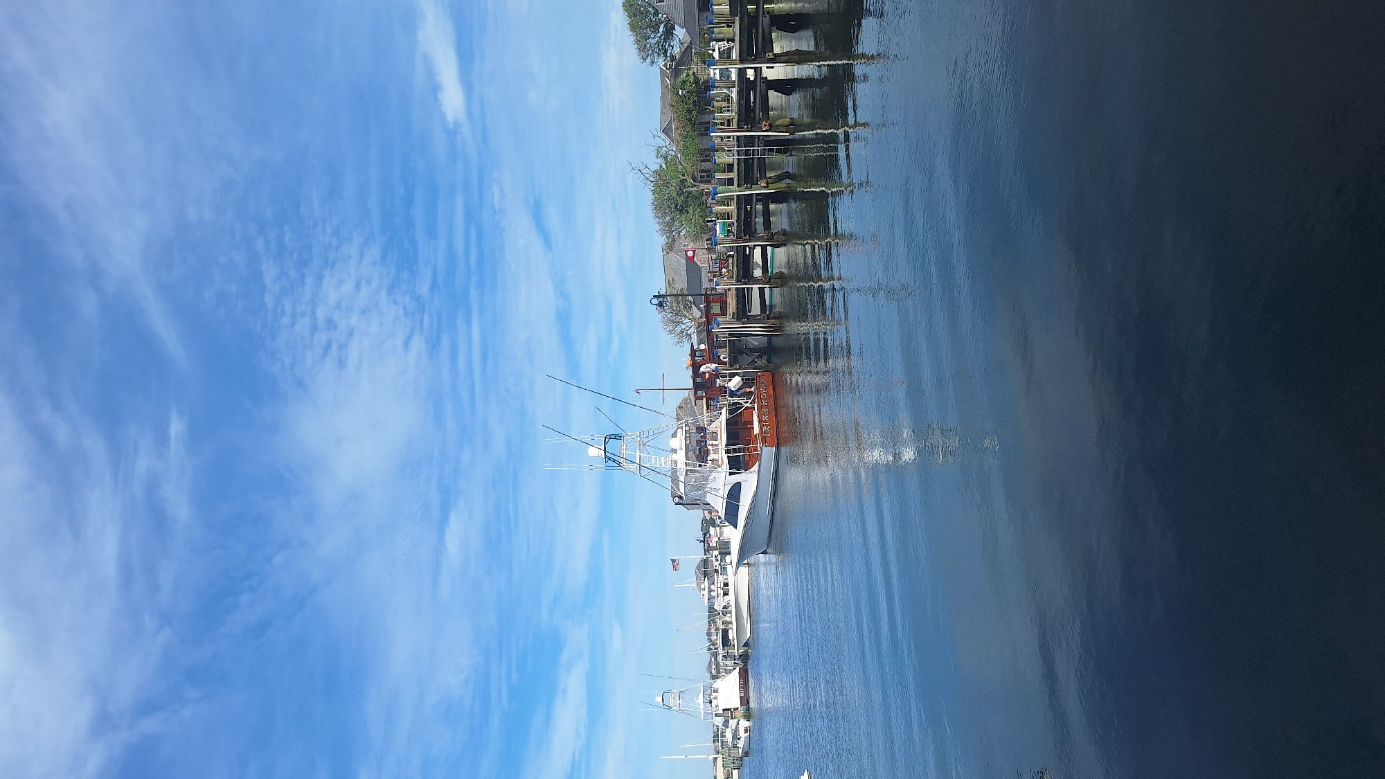 This is a full-frame, eye-level, outdoor, vertical, natural light photograph taken from a waterfront perspective. The scene depicts a harbor with several boats docked at a wooden pier. In the foreground, the deep blue water is calm with slight ripples, reflecting the sky and the boats. The sky is a bright blue with scattered white clouds, some of which are wispy and elongated.
Dominating the mid-ground is a large, white sportfishing boat with a prominent tower structure. The boat has a reddish-brown hull with "IRISH ROVER" inscribed on its side in white lettering. Several other boats, also docked, are visible to the left and behind this main vessel, including a white boat with "RIFFED" visible on its hull. The pier itself is made of weathered wood pilings and planks, with ladders visible along its edge. A few people can be seen on the pier and on one of the boats, engaged in what appears to be routine activity.
In the background, beyond the pier and boats, are several buildings with a New England architectural style, characterized by their muted colors and pitched roofs. Trees with green foliage are also present, suggesting a temperate climate. The overall atmosphere is tranquil and suggests a leisurely day in a coastal town. The image location is the town of Nantucket, Massachusetts, United States of America, specifically likely the Nantucket Boat Basin or a similar harbor area.