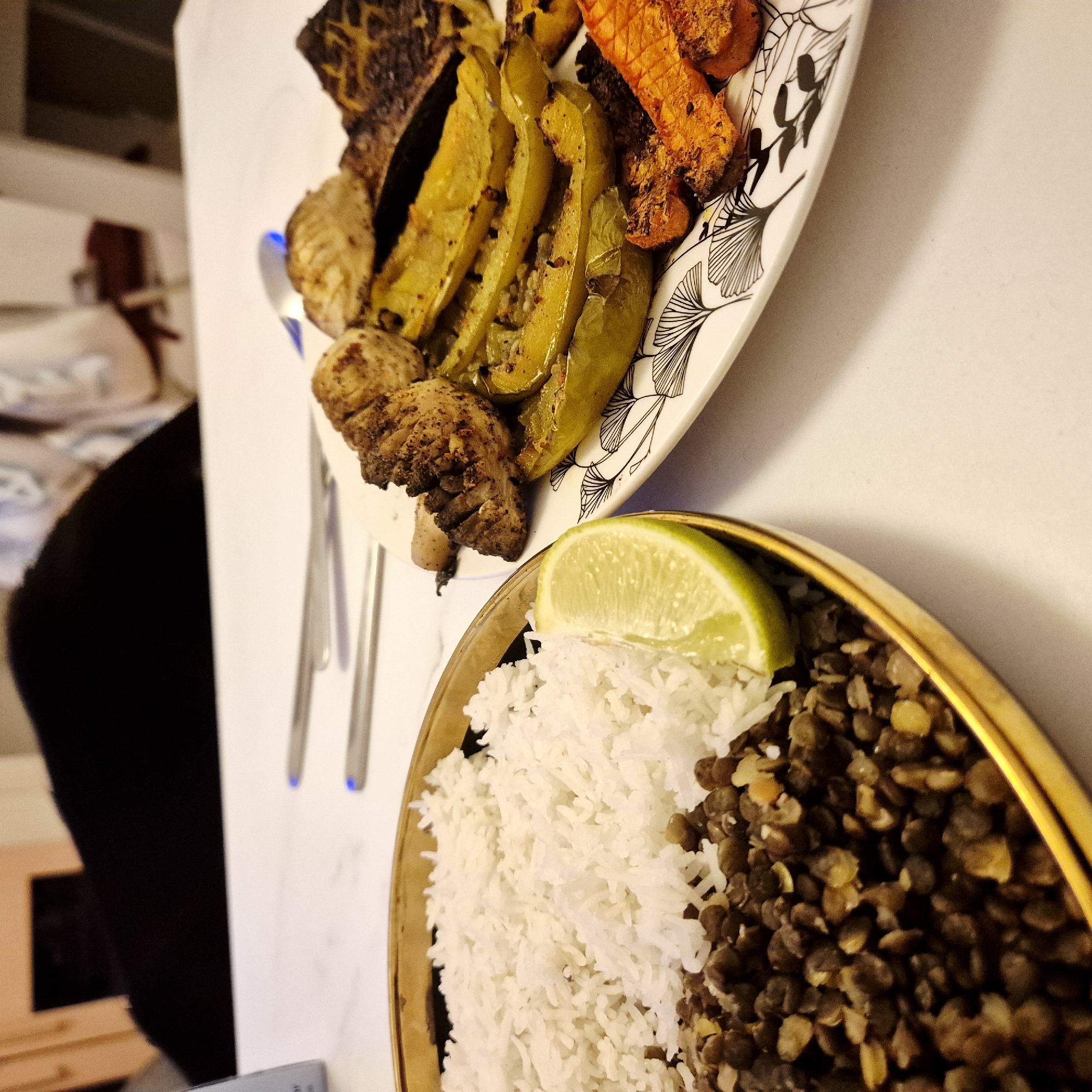 The image captures two plates of food on a white surface. In the top plate, there are grilled vegetables including bell peppers, mushrooms, and carrots. The plate has a leaf design on the edge. Next to this plate there are two sets of chopsticks. The bottom plate has white rice, lentils, and a lime wedge. The lower plate has a gold rim. The location is likely indoors. The scene does not appear to be in Strasbourg, France. The objects present, specifically the food, plate designs, and chopsticks suggest the location might be in Asia.
