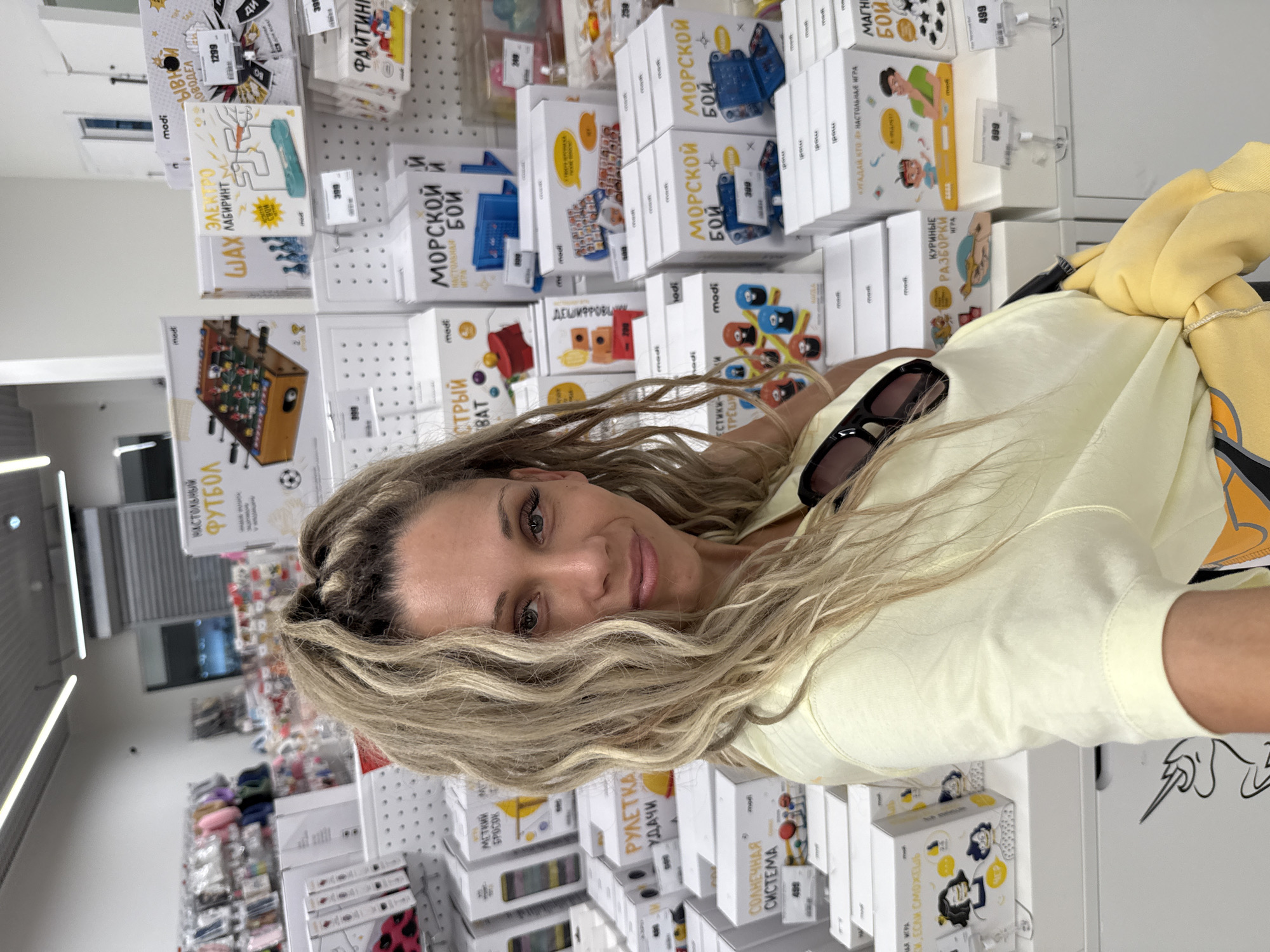 A woman with blonde, curly, pulled-back hair is taking a selfie in a store. She is wearing a pale yellow t-shirt and sunglasses are pushed up into her hair. The background is filled with shelves of boxed toys. The boxes are mostly white with colorful graphics and Russian text. Some of the titles visible on the boxes include "Настольный Футбол" (Table Football), "Электрический Лабиринт" (Electric Maze), "Морской Бой" (Sea Battle), "Рулетка Удачи" (Roulette of Fortune), "Солнечная Система" (Solar System), and "Куриные Разборки" (Chicken Troubles). The store appears to be well-lit with fluorescent lighting. The overall scene suggests a toy store environment.