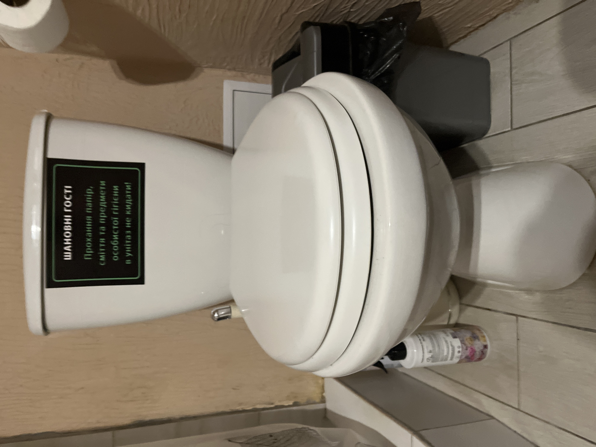 A white ceramic toilet is centrally positioned in the frame. A black and green sign is affixed to the tank, displaying text in Ukrainian. To the right of the toilet, a grey trash bin with a black plastic liner is visible. A bottle with colorful contents stands to the left of the toilet base. The floor is tiled with light-colored squares. The walls appear to be a textured beige. The scene is indoors, likely a bathroom.