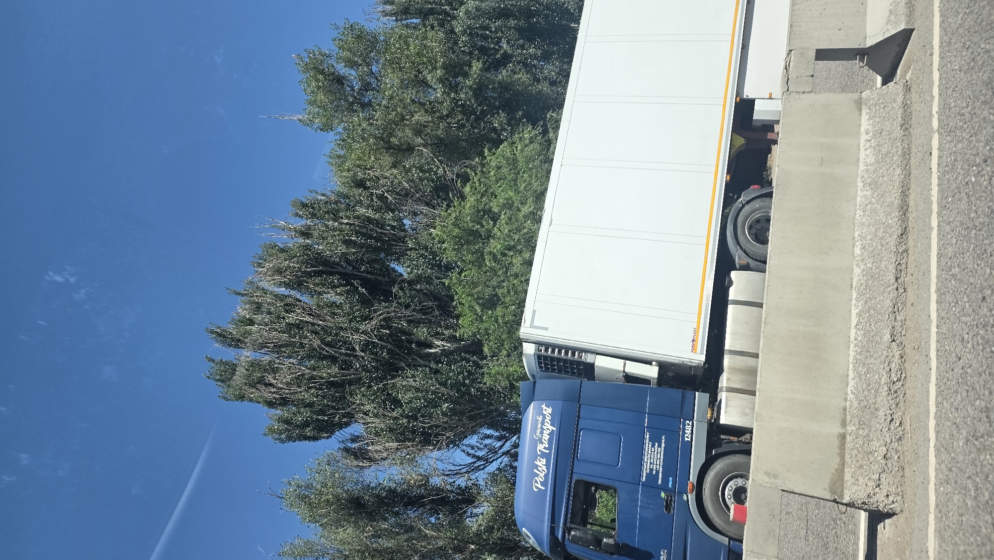 A blue semi-truck with a white trailer drives on a highway with trees and a blue sky in the background. The truck has "Polski Transport" written on the side of the cab. The trailer is a refrigerated unit. The truck is driving behind a concrete barrier that separates the lanes of traffic. The image appears to be taken from another vehicle on the highway.