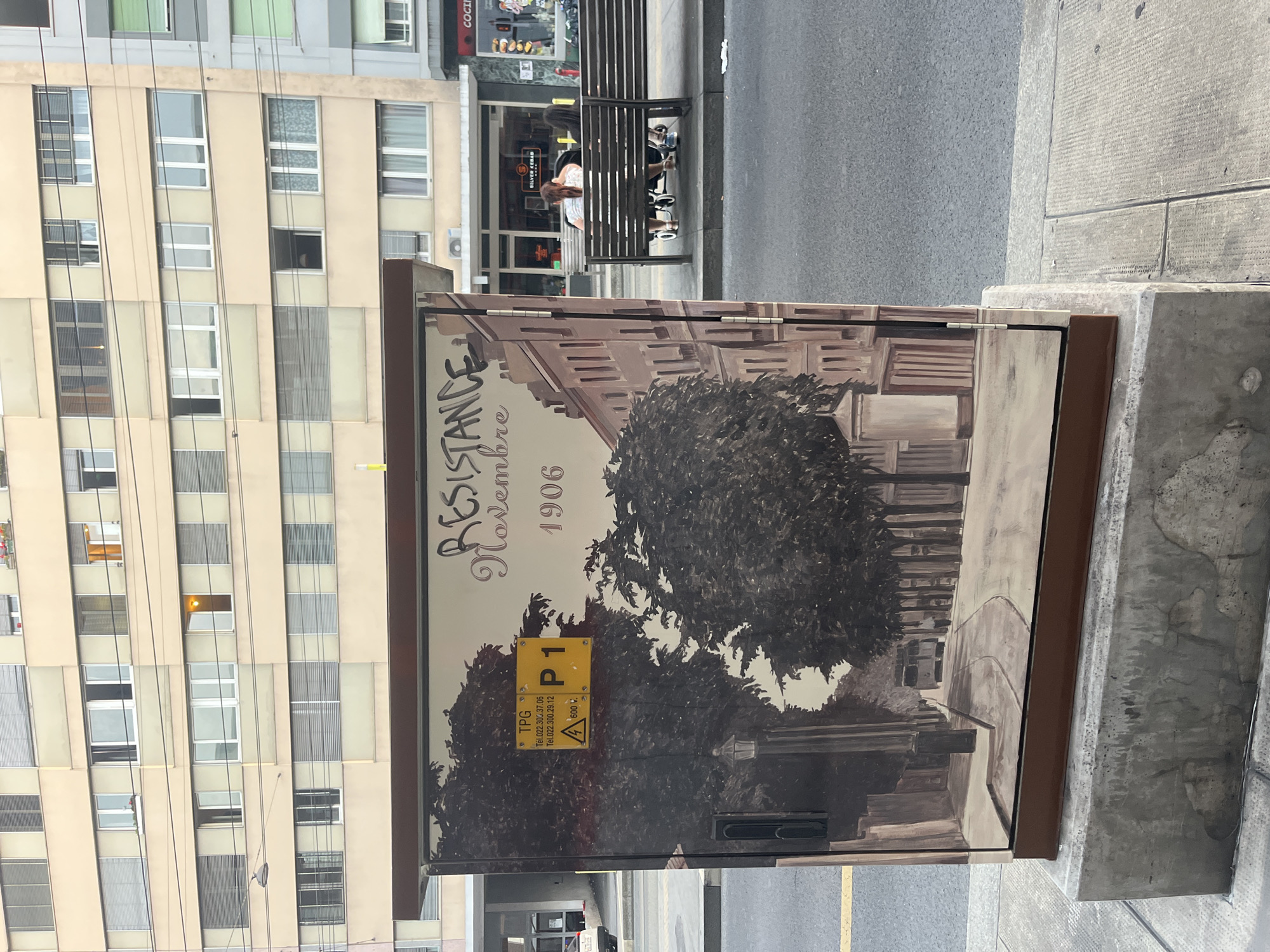 The image depicts a street scene in Geneva, Switzerland, focused on a large, decorated electrical cabinet. The cabinet features a mural depicting a European city street with trees, buildings, and a tram in the distance. Text on the mural reads "RESISTANCE Novembre 1906." A yellow warning sign with "TPG" and phone numbers is affixed to the cabinet, along with a "P1" designation and a high-voltage warning symbol. To the right of the cabinet, a public bench is occupied by two women and a baby stroller. In the background, a multi-story building with numerous windows lines the street, and shops are visible. The environment appears to be an urban setting with paved sidewalks and a street.