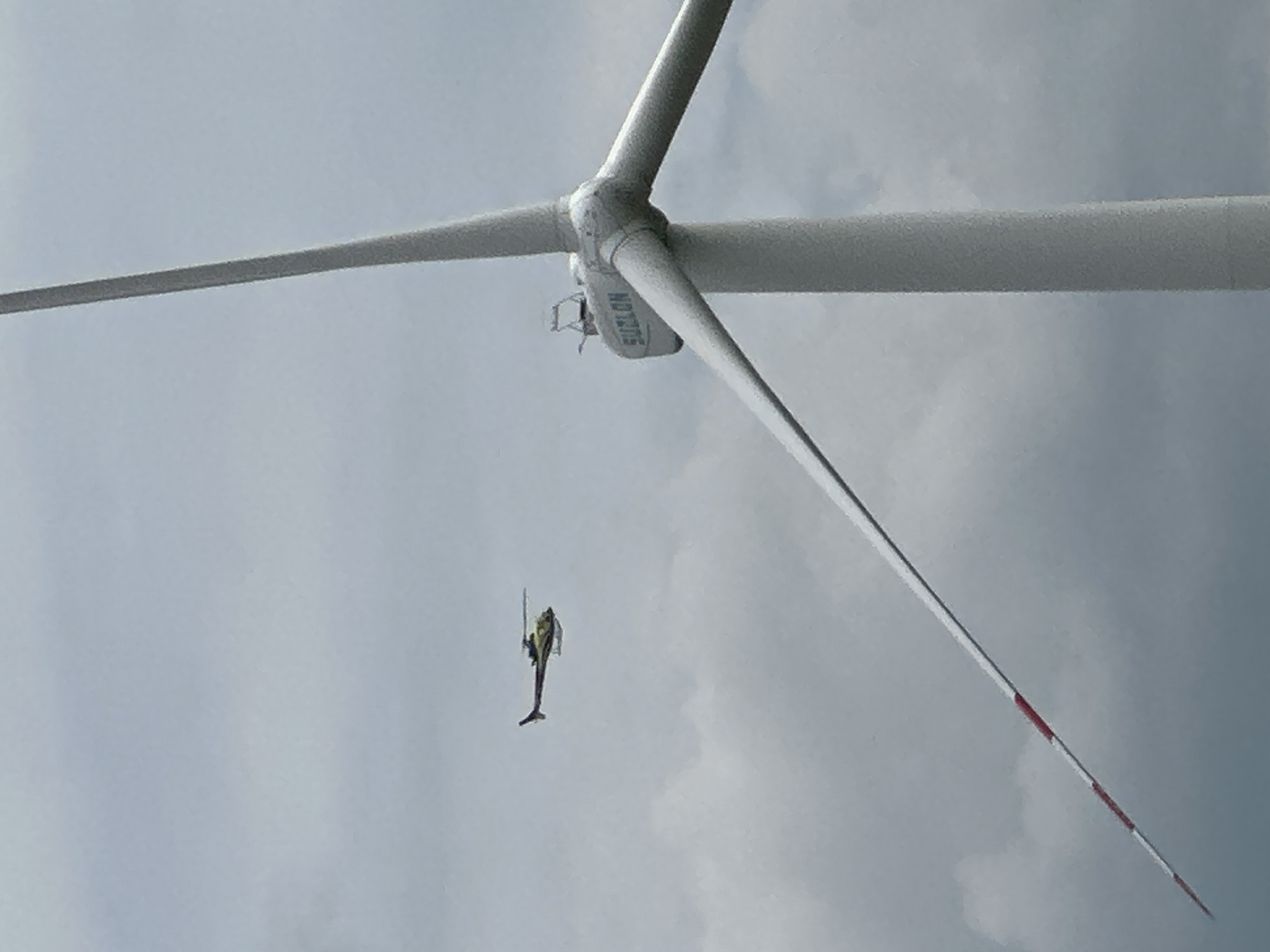 A helicopter is flying in the sky near a wind turbine. The helicopter is small and yellow and appears to be in flight. The wind turbine is tall and white, with three blades. One of the blades is in the foreground and has red and white markings on it. The sky is cloudy and overcast. The scene is set in the city of Korchivka, Ukraine.