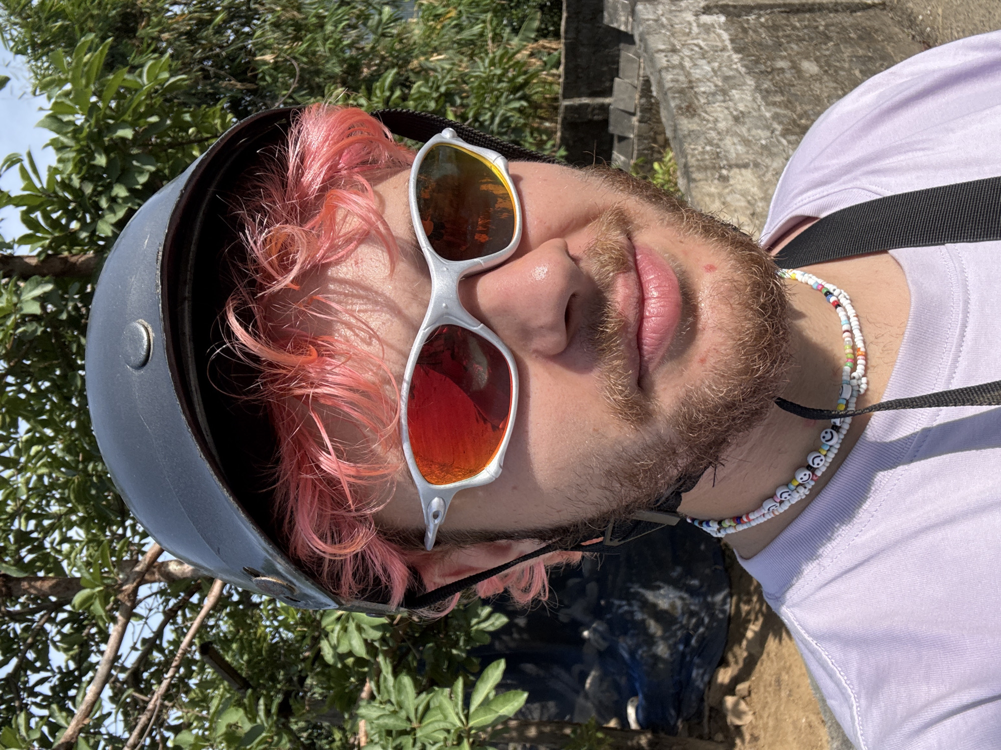 A close-up shot of a person with pink hair wearing a black helmet and silver-framed sunglasses with reflective lenses. The person has a beard and is wearing a white T-shirt with a beaded necklace. The background shows greenery, a stone structure, and glimpses of the sky. The location is in Đà Nẵng, Vietnam.