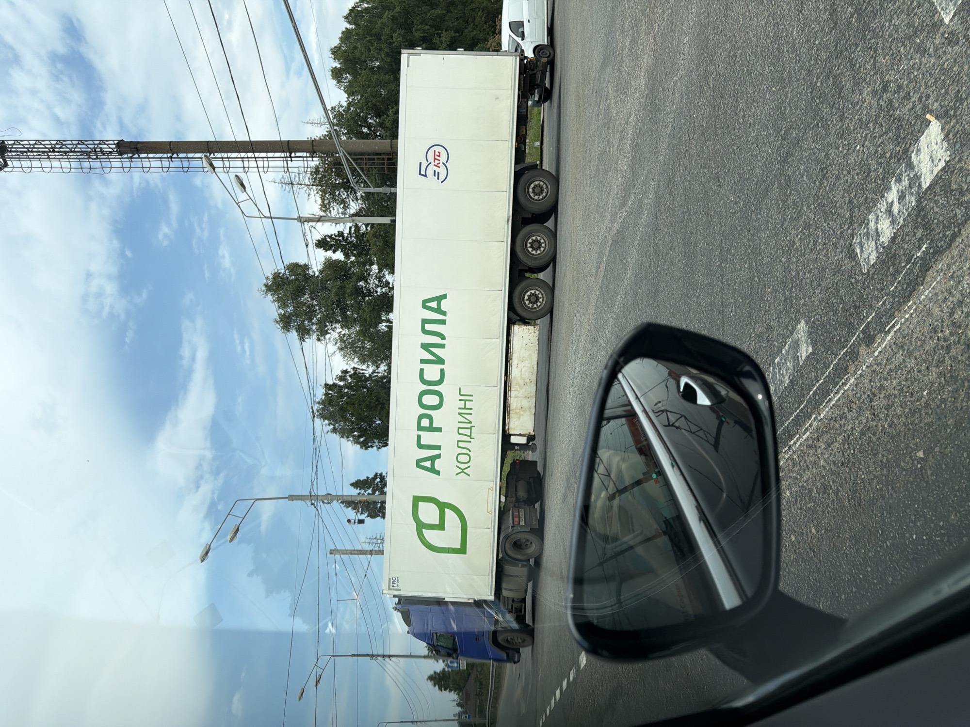 This image, taken in Radumlya, Russia, captures a view from a vehicle's side mirror reflecting the road and sky, while the main scene shows a large white semi-trailer truck with "АГРОСИЛА" and "ХОЛДИНГ" written in green on its side. The truck has a blue cab and is traveling on a paved road. A white van is visible behind the truck, also on the road. The background features trees, power lines, and a concrete tower with a ladder. The sky is partly cloudy. The road has white lane markings.