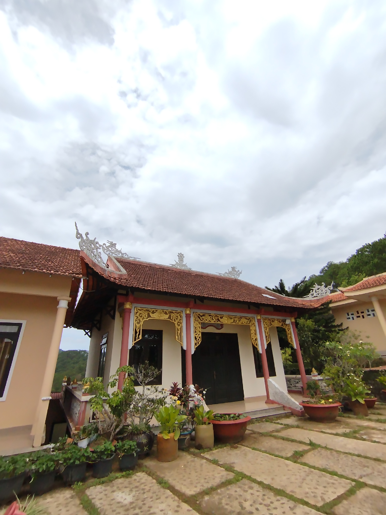 The image shows a low-angle perspective of a building with traditional Vietnamese architectural features in Da Lat, Vietnam. The main building in view is a single-story structure with a red tile roof and ornate white and gold trim along the eaves and gables. The walls of the building are painted white, and it has dark-colored doors and windows. The building is situated on a paved terrace, and there are numerous potted plants with various flowers and greenery arranged around the base of the building and along the terrace. A portion of another building is visible on the left side of the image, featuring a similar architectural style and color scheme. The sky is overcast with dense gray clouds. The surroundings indicate a hillside location, with trees visible in the background. The photo appears to have been taken during the daytime with even lighting.