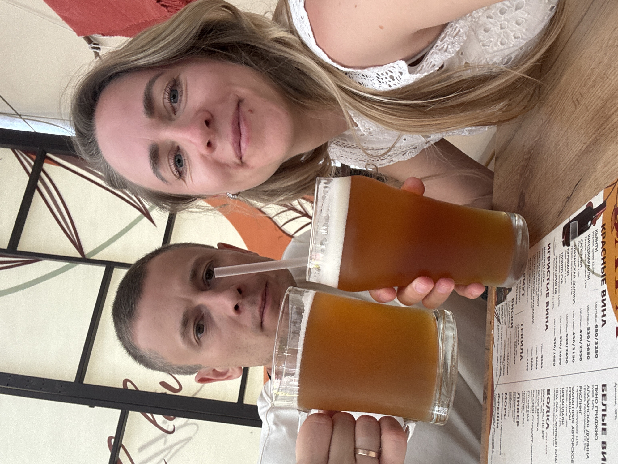 This is a selfie taken by two people holding glasses of beer, suggesting they are at a bar or restaurant. The woman in the foreground has blonde hair and is wearing a white lace top. The man in the background has short, grey hair. The background shows a table with a menu and what appears to be some decorative elements of the establishment. The lighting suggests it is daytime. The menu is in Russian and lists various alcoholic beverages, including wines, vodkas, and whiskies. The scene appears casual and relaxed, with the subjects smiling and holding their drinks up, possibly for a toast. The location is identified as Chaikovsky, Russia.