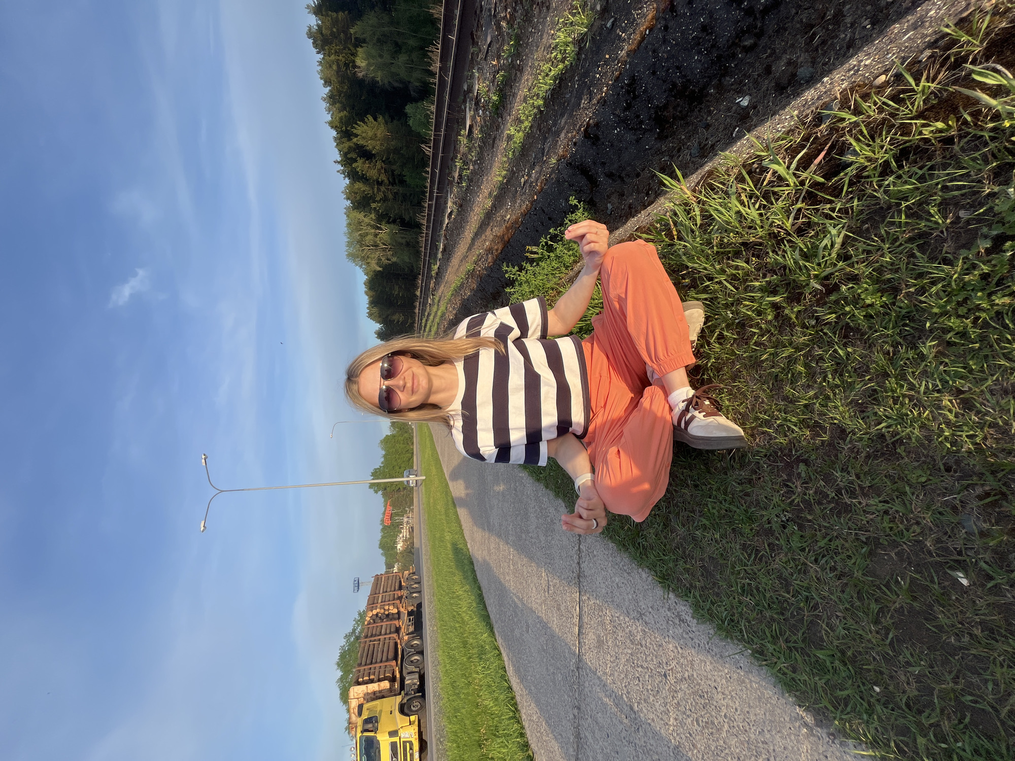 A woman is sitting cross-legged on the grass beside a sidewalk. She has long blonde hair and is wearing sunglasses, a black and white striped shirt, and orange pants. She is positioned near railroad tracks. The location is Chaikovsky, Russia, suggested by a logging truck loaded with logs visible nearby, typical of the region. The background includes trees, a clear sky, and streetlights.