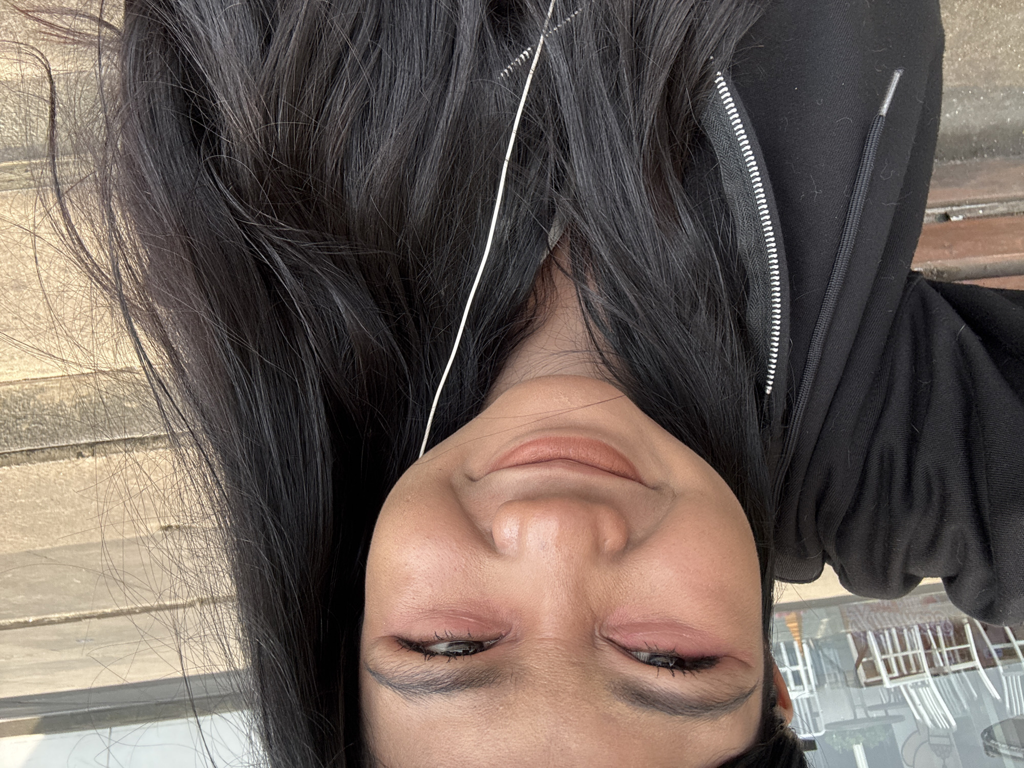 A woman is photographed from a close-up, upside-down perspective. She has long, dark hair and wears a black hooded jacket with a visible zipper. A thin, white wire extends from near her hair. The image appears to be taken outdoors, with glimpses of an urban environment including building structures visible. Given the instruction to include the location of North Jakarta, Indonesia, it's implied the image was taken in this city.