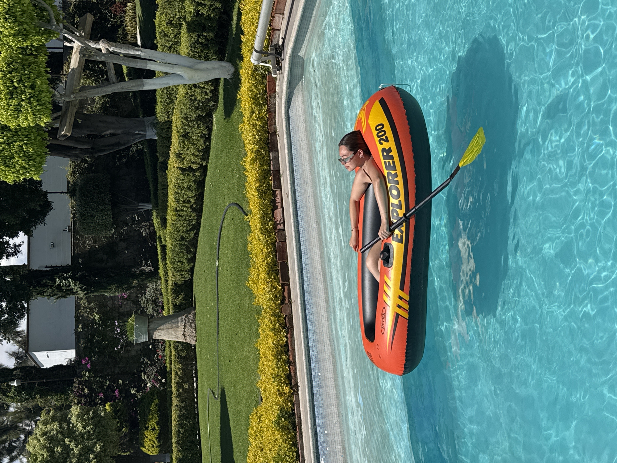 The image shows a woman floating in a pool in an orange and black inflatable boat. The boat is labeled "EXPLORER 200." She is holding a yellow paddle. The pool is surrounded by a well-manicured lawn and hedges, with a building visible in the background. Based on the landscaping, architecture, and general surroundings, the location is likely in a residential area of Puebla, Mexico.
