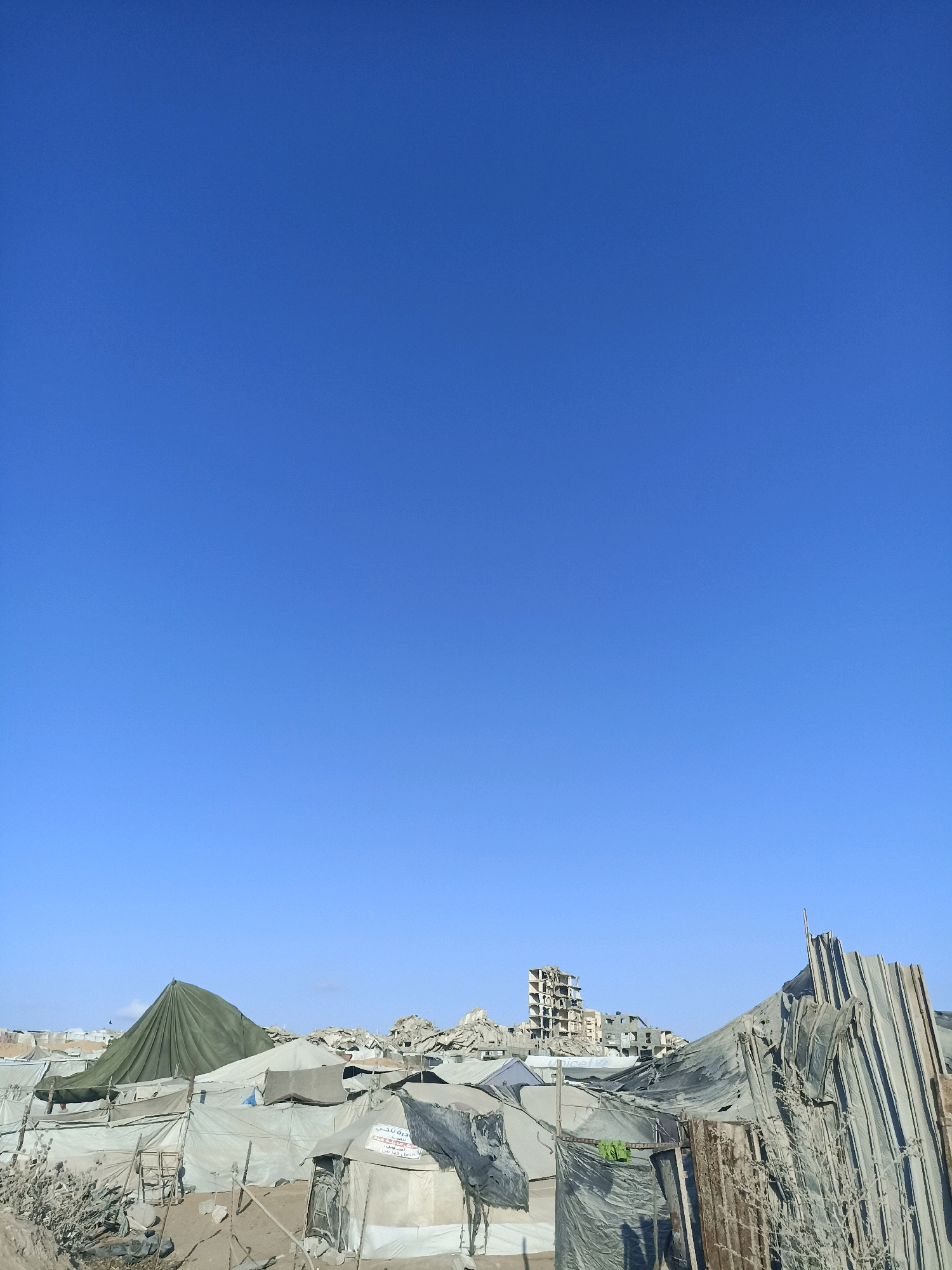The image depicts a scene of widespread destruction in Gaza City, Palestinian Territories, under a clear blue sky. In the foreground, makeshift tents and shelters constructed from tarpaulins and salvaged materials are visible, indicating a displacement of people. Some tents are erected over debris and rubble, suggesting they are temporary living spaces. A large, olive-green conical tent stands prominently among the other shelters.
In the background, the skeletal remains of damaged buildings are scattered across the landscape, with some structures appearing to be partially or completely destroyed. One damaged building in the mid-ground has multiple floors exposed. Piles of rubble and debris are visible throughout the scene, further emphasizing the extent of the damage. The environment appears arid and dusty. No individuals are clearly visible in the image. The overall impression is one of devastation and the struggle for survival in a war-torn urban environment.