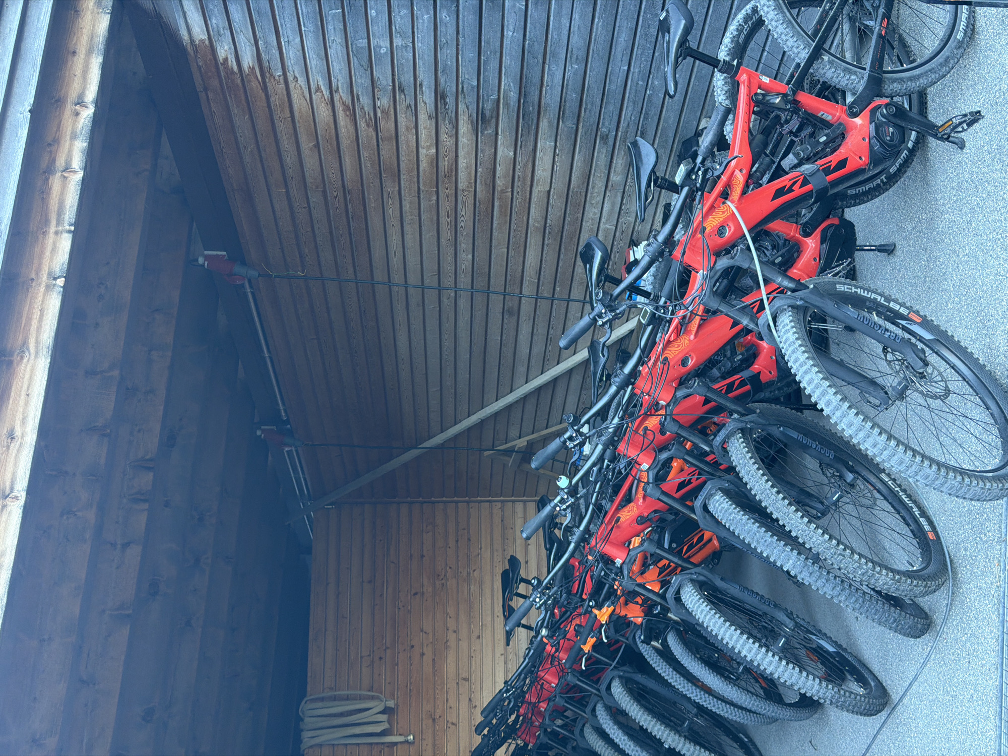 A row of bright orange electric mountain bikes is parked in a covered wooden structure. The bikes are lined up closely, facing towards the viewer. Each bike has a full suspension frame, knobby tires, and a visible battery pack on the down tube. The structure appears to be open on one side, revealing the bikes, and has a wooden plank wall and a wooden ceiling. A coiled green hose is visible on the left side of the structure, attached to the wooden wall. A power cord with a red plug is hanging from the ceiling, suggesting the bikes may be charging. The ground is paved with asphalt. The scene is well-lit, with natural light illuminating the bikes and the interior of the structure. The overall impression is that of a bike rental or storage area for recreational vehicles.
