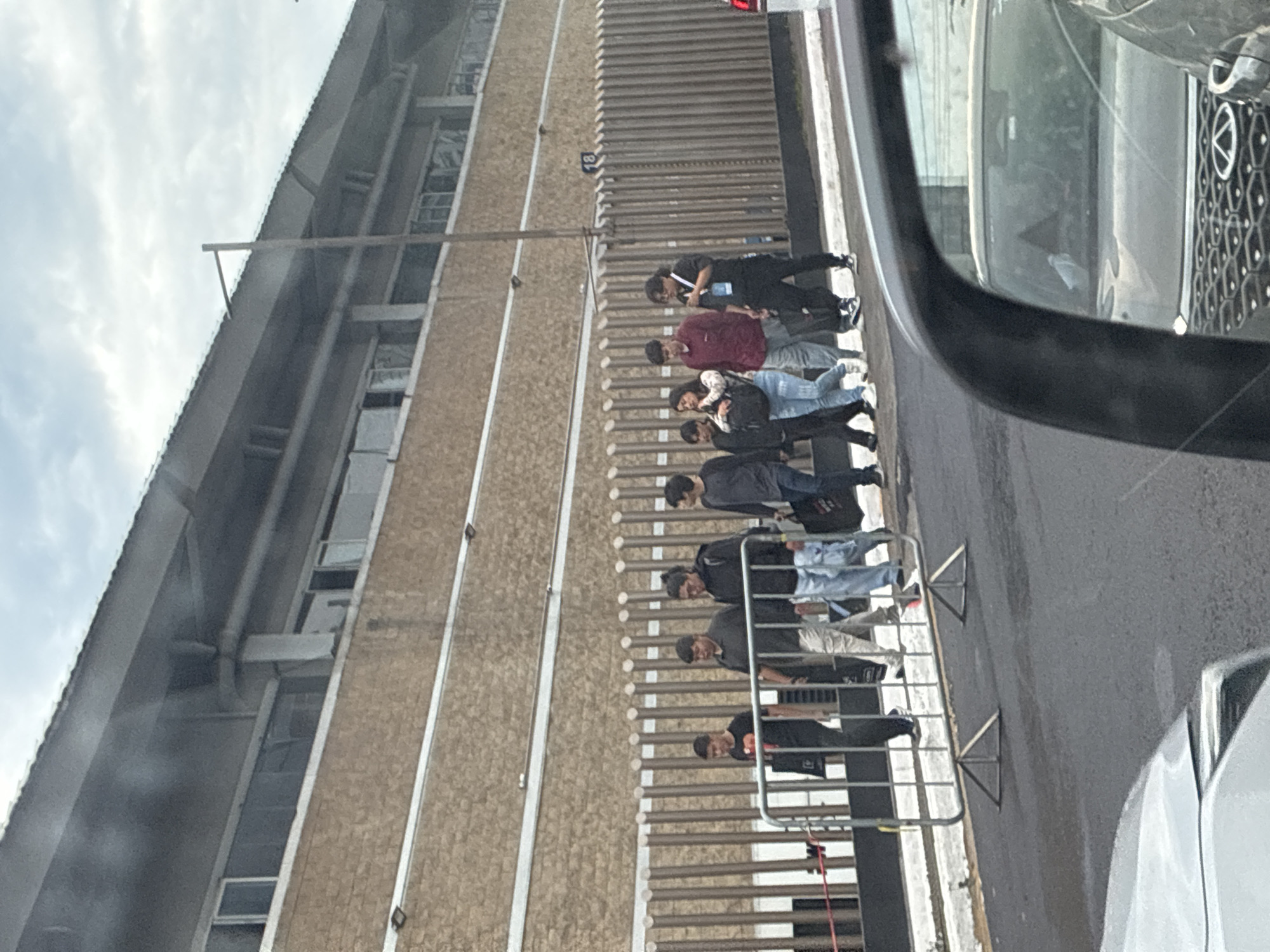 Scene captured in Mexico City, Mexico. A line of approximately eight individuals, including both men and women, standing on a sidewalk next to a building with multiple stories. They are standing behind a series of metal barriers. Some are looking at their mobile phones. There is a car or other vehicle partially visible in the right side of the frame, showing the side mirror and portions of the front. The weather appears overcast.
