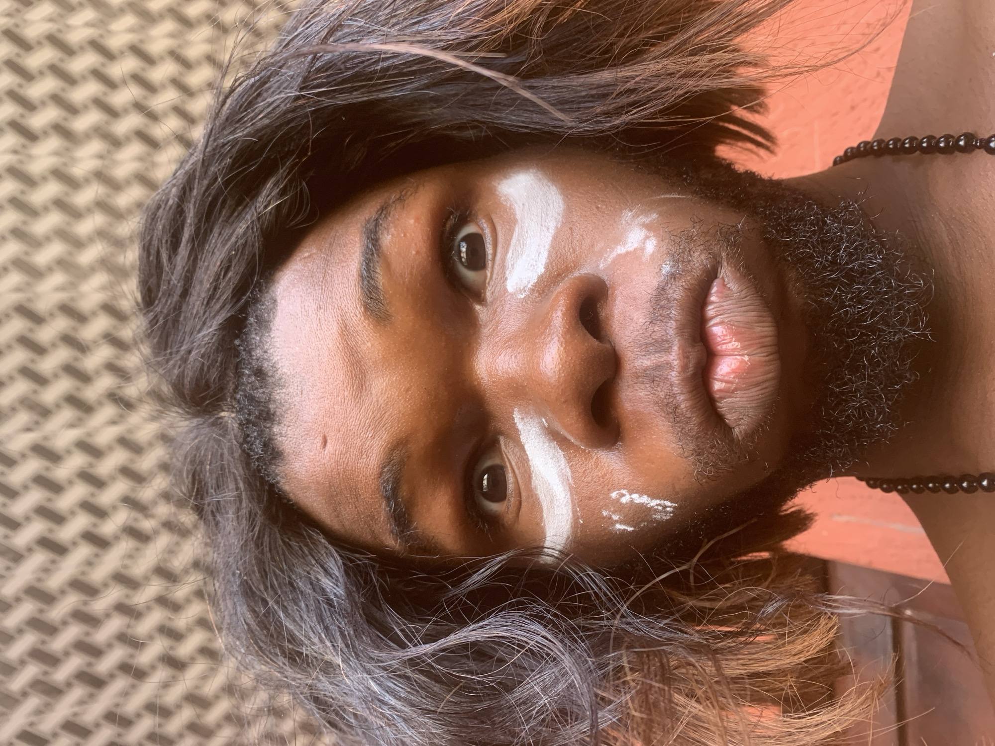 A close-up portrait captures a person with dark skin, positioned with their head tilted, looking directly into the camera. The individual has long, dark hair, with hints of reddish-brown strands, appearing styled or naturally voluminous around their face. They sport a dark, short beard or stubble on their chin and cheeks.
Prominent white markings are painted on the face: a broad stroke across the forehead, another under the left eye (viewer's right), and smaller, scattered marks on the right cheek (viewer's left). The person's eyes are dark, and their lips are full. A black beaded necklace is visible around their neck on the right side of the frame.
The background features a textured surface with a repeating geometric pattern in light brown and beige tones, resembling woven material or tiles. Behind the person's right shoulder and neck, there is a plain, warm orange or reddish surface. The lighting appears natural, casting subtle highlights on the skin and hair. The expression is neutral and direct, suggesting a contemplative or engaging gaze. This image, taken in Nkpor, Nigeria, could be a portrait showcasing cultural adornment, a performance, or an artistic expression. There is no visible text in the image.