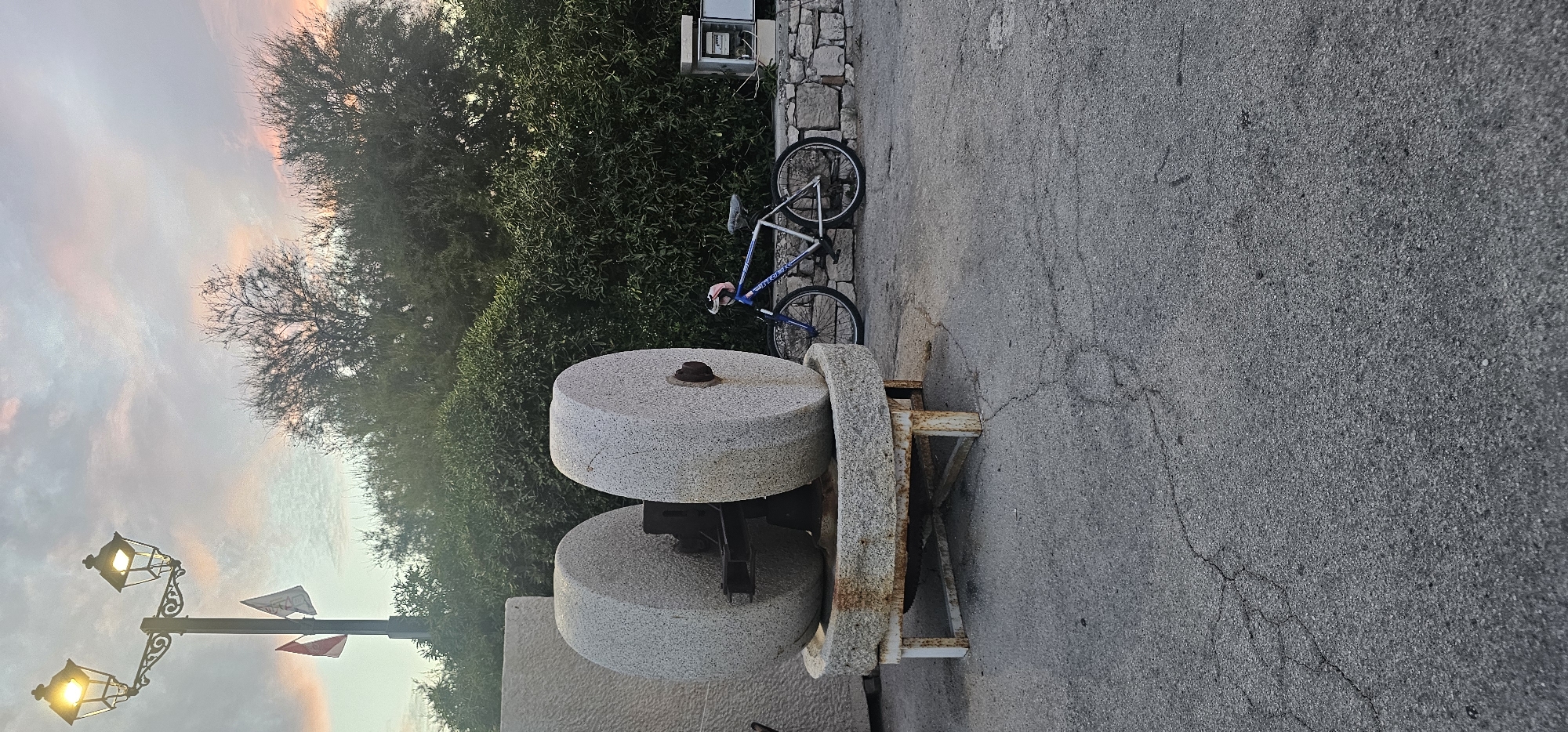 In the city of Supetar, Croatia, at dusk, a large stone olive press stands in the foreground. The press is composed of two massive, weathered granite wheels resting on a circular stone base, which is supported by a rusted metal frame. To the right of the olive press, a blue bicycle with a pink handlebar accessory leans against a low stone wall. Behind the wall, lush green bushes form a dense backdrop. A lamppost with two illuminated lamps stands to the left of the frame, with two small flags attached to its pole. The sky above is filled with pastel-colored clouds, indicating the setting sun. The ground is paved with cracked asphalt. A small, open electrical box is visible on the stone wall to the right of the bicycle.