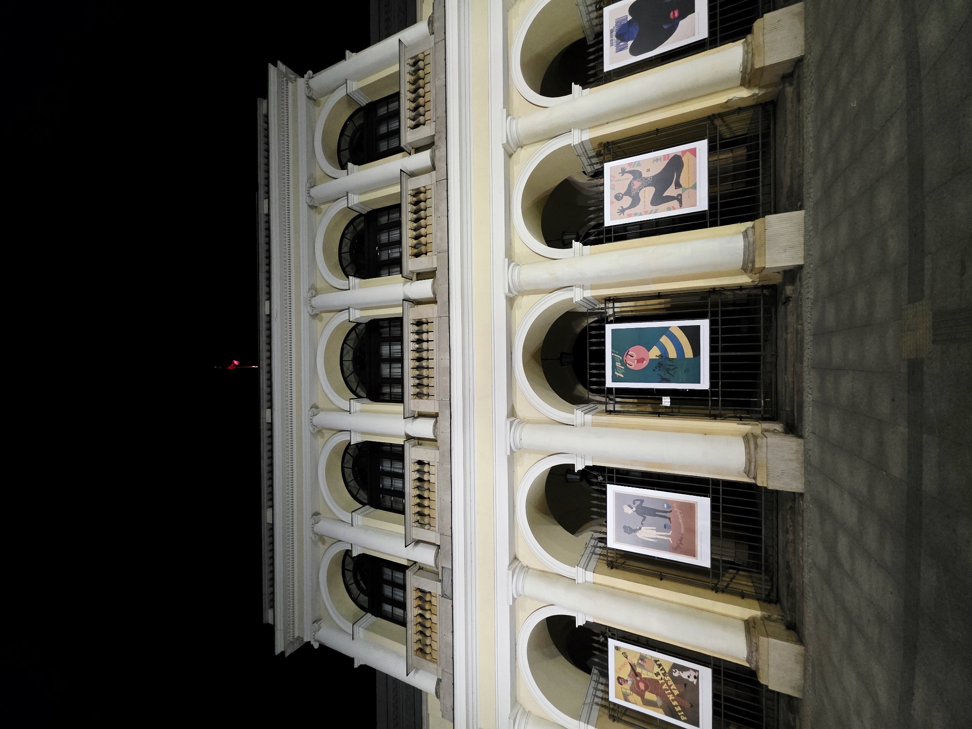 The image shows the facade of a cream-colored building with arched windows and pillars at night. Several posters featuring colorful, stylized illustrations are displayed on metal fencing in front of the building's lower level. The building's architecture suggests a historical or cultural institution. The presence of posters indicates an event or exhibition is taking place. The sky is black, suggesting it is nighttime. A small red and black flag is visible on the rooftop. The image is taken from a low angle, looking upwards at the building.
The building is the National Theatre in Warsaw, Poland.