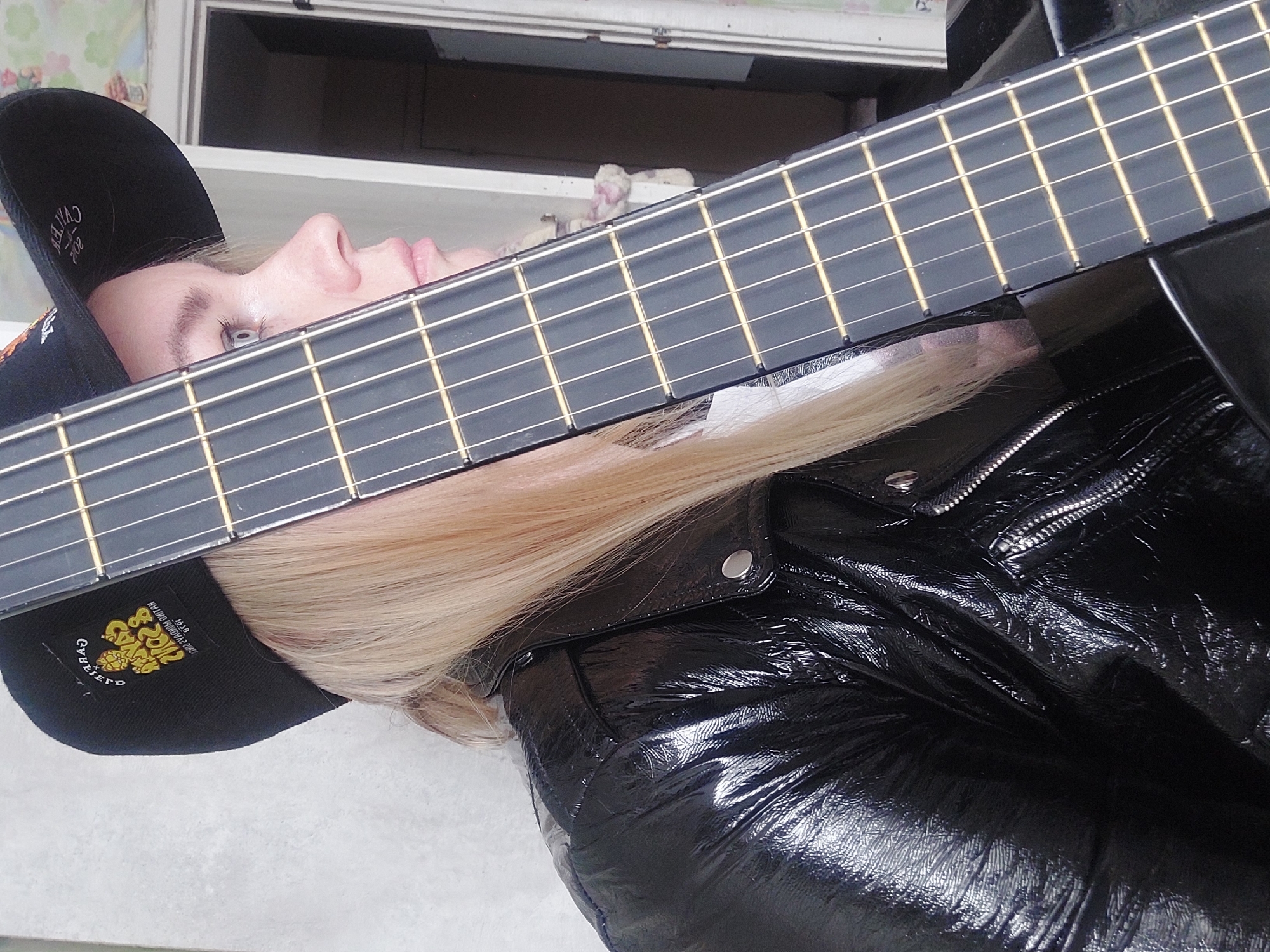 A female with blond hair is wearing a black baseball cap with a logo and a shiny black jacket. She is lying down while holding a guitar in front of her face. The location is likely in Chaikovsky, Russia, but this is inferred based on the image and is not directly depicted. The background includes a window and a wall with a floral pattern.