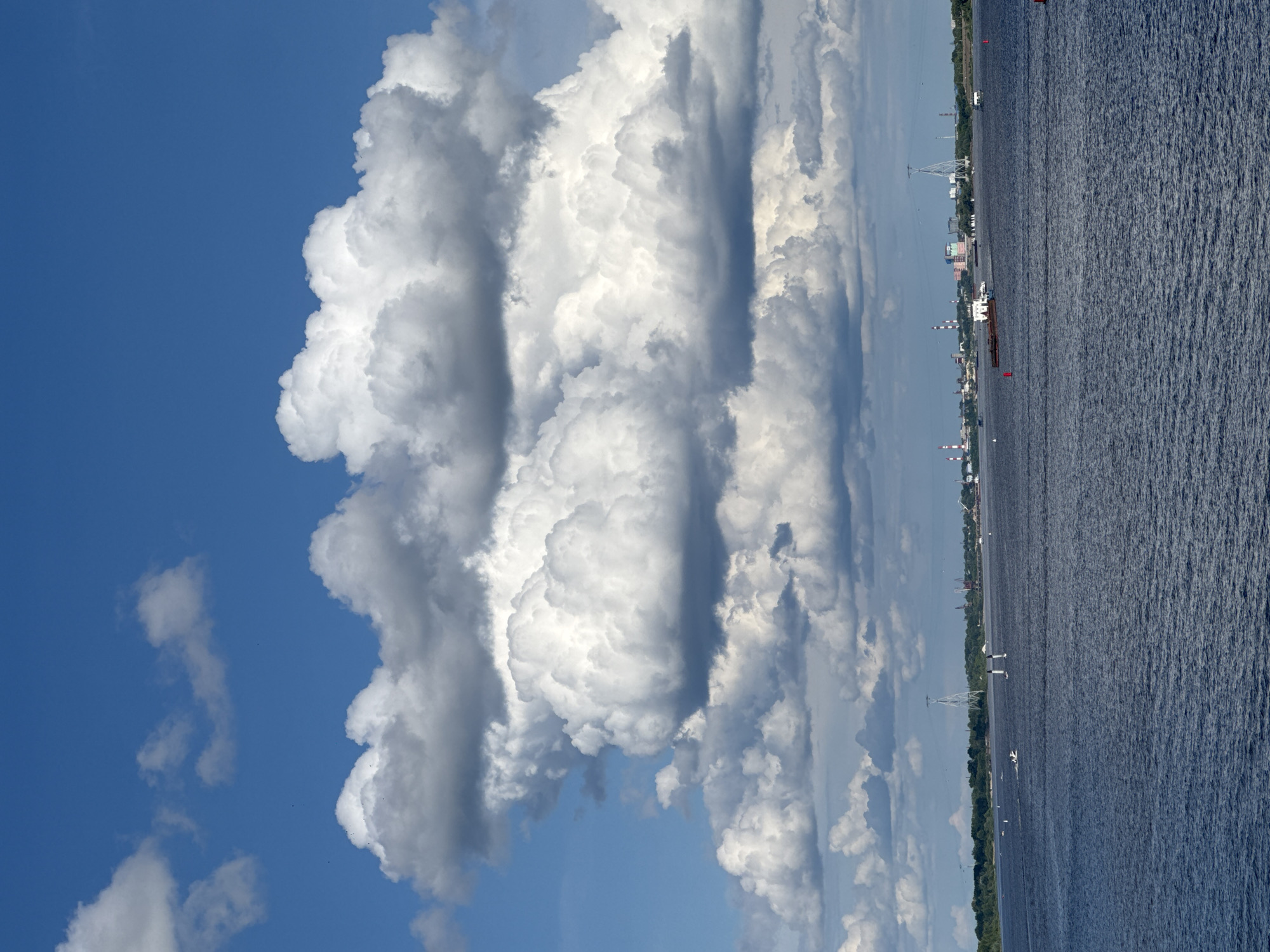 The image depicts a wide, dark blue river with rippling water under a bright blue sky filled with dramatic, voluminous white and gray cumulus clouds. On the horizon, a distant city skyline is visible, characterized by industrial structures like smokestacks and cranes, along with buildings. Several boats and a barge are scattered across the water, indicating maritime activity. The overall scene suggests a view from a waterfront in Nizhny Novgorod, Russia, possibly during a sunny day with developing cloud formations.