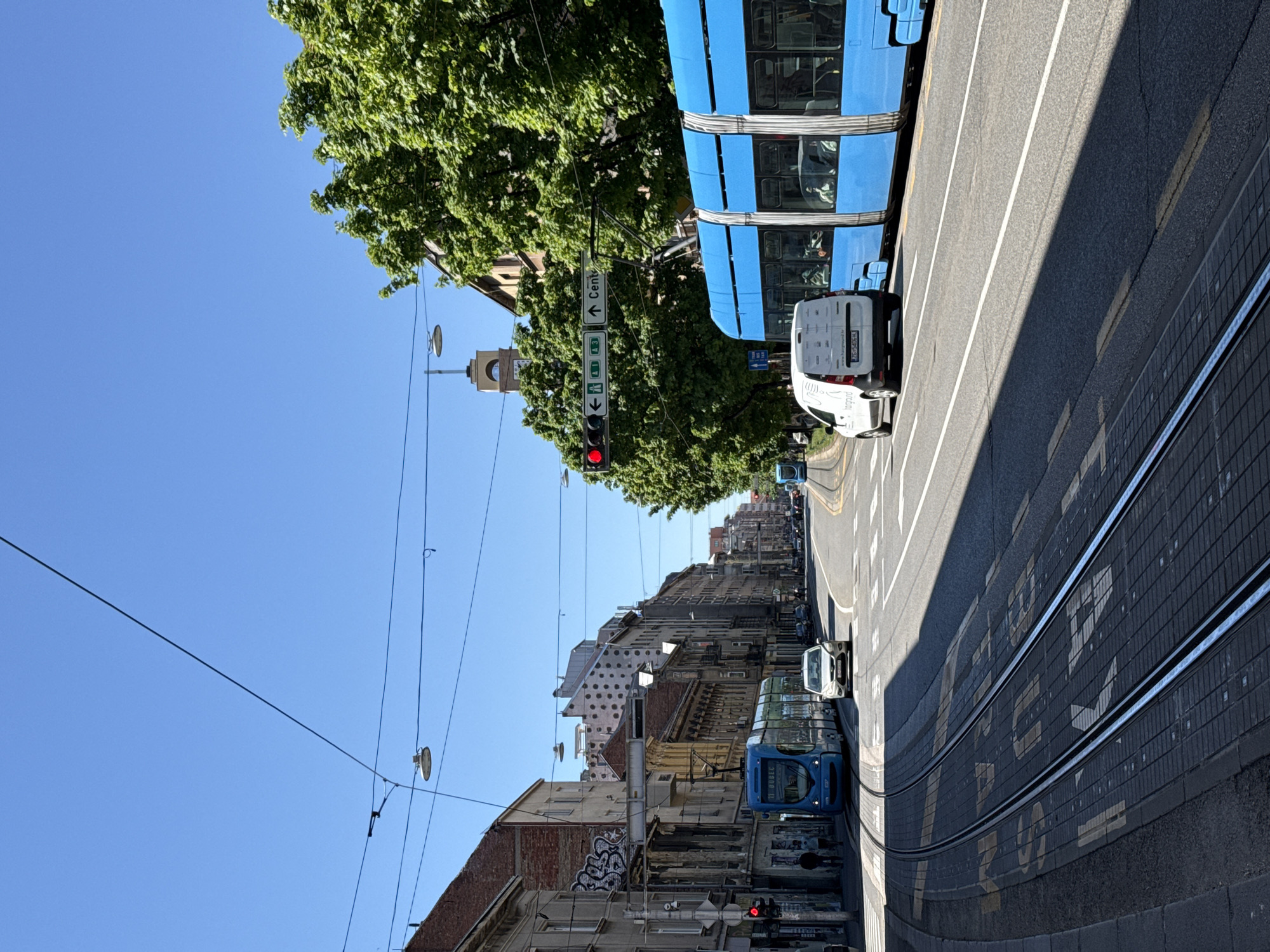This image captures a vibrant, sunny day on a city street in Zagreb, Croatia, viewed from a slightly elevated perspective looking down the thoroughfare. The scene is dominated by a wide road with tram tracks running parallel, receding into the distance.
On the right side, a modern, bright blue tram is prominent, either stationary or moving slowly. Its windows reflect the bright sky and surrounding greenery. Behind it, a white delivery van is parked or moving. Another blue tram is visible further down the street on the left side of the tracks. The road features clear white lane markings and painted text "P + R".
To the left, a line of multi-story buildings with varied architecture stretches along the street, some displaying graffiti. Overhead, a dense canopy of green trees provides shade, casting sharp, elongated shadows across the road, indicating a strong sunlight, likely midday or early afternoon. The sky above is a clear, cloudless blue. Numerous overhead power lines for the trams and streetlights crisscross the upper portion of the frame.
A red traffic light is visible in the mid-ground, alongside a street sign pointing "↑ Centar" (Center) and "↓ A3", "↓ A1". The tram has "11 DUBEC" displayed, likely its route and destination. The white van bears the text "www.hargraund.hr" and "hargraund", with a Croatian license plate "ZG 553-EK". In the background, rising above the buildings and trees, a distinct clock tower with a dome is visible. The overall scene suggests a busy urban environment with active public transport and vehicular traffic.