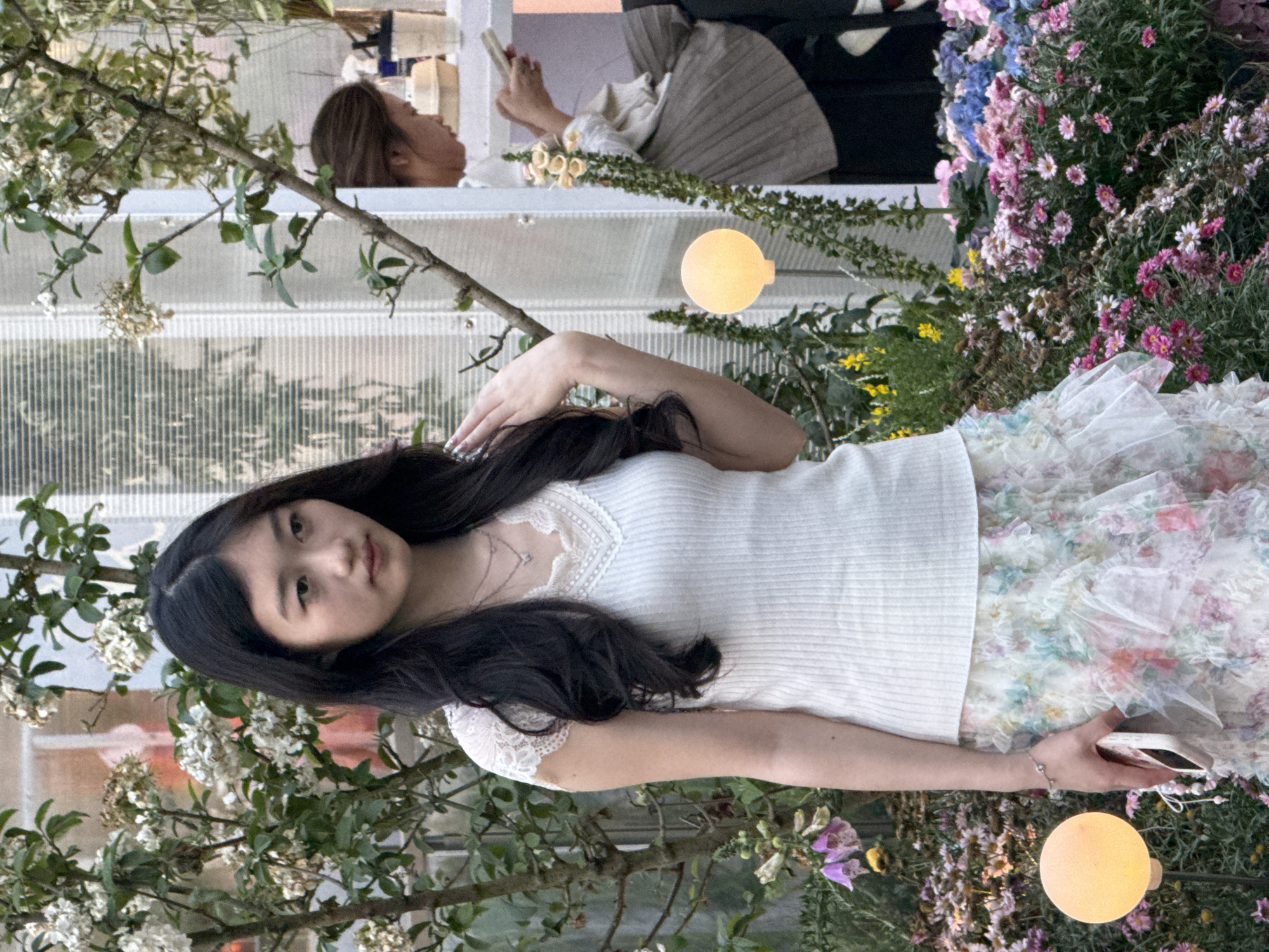 The image shows a young Asian woman posing among foliage and flowers, likely in an indoor setting given the window-like structures in the background. She is wearing a white top and a floral patterned skirt, holding a phone in her right hand and raising her left hand towards her head. There are illuminated spherical lights placed in the surrounding greenery. Another person is visible in the background looking at a phone. The location cannot be confirmed as Liangjiang, China, from visual information.