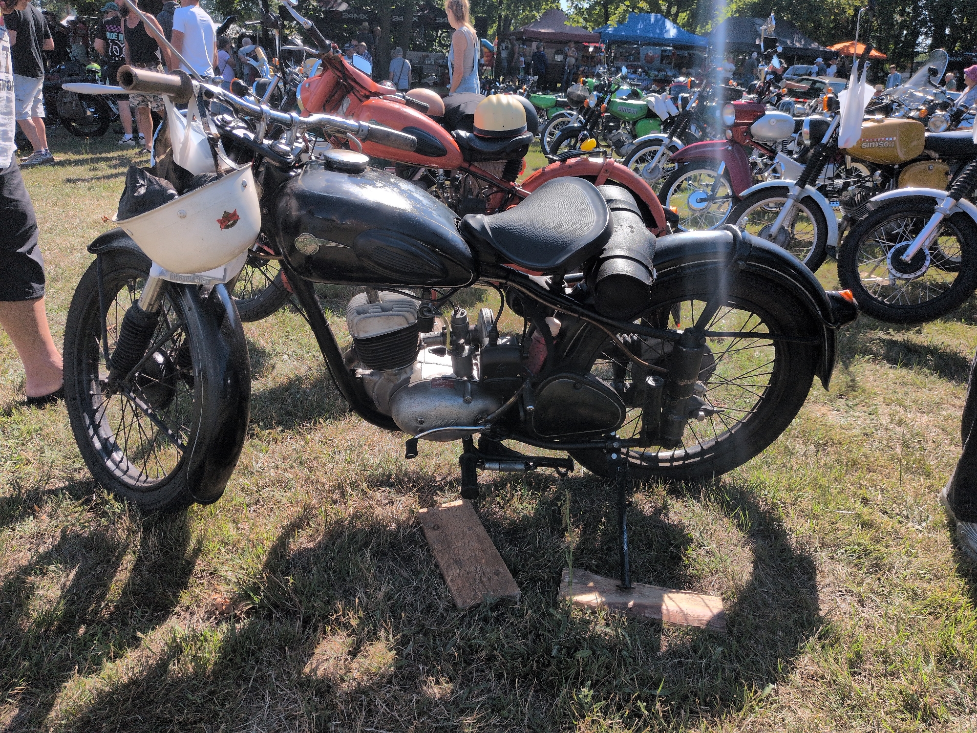 The image captures an outdoor scene at the 2023 motorcycle meeting in Hohenferchesar, Germany. The foreground is dominated by a vintage black motorcycle with a white helmet attached to its handlebars. The motorcycle features a black leather seat, a chrome exhaust pipe, and spoked wheels. The scene is set on a grassy field, with shadows cast by the sunlight suggesting it is daytime. In the background, several other motorcycles and people are visible, along with tents and trees, indicating a gathering or event. The overall atmosphere appears to be relaxed and focused on the exhibition of classic motorcycles.