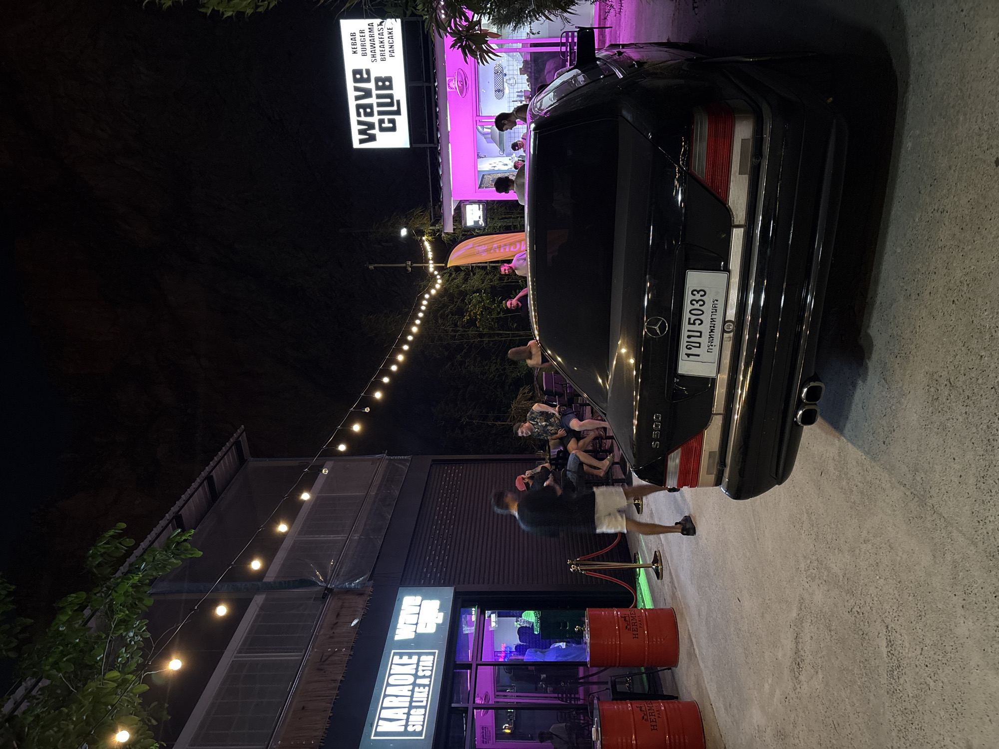 A night scene in Baan Bang Rak, Thailand, shows a black Mercedes S500 partially blocking a sidewalk. The car's license plate reads "1ขบ 5033 กรุงเทพมหานคร". Behind the car, people are gathered at tables outside a karaoke bar named "KARAOKE Wave". A sign above reads "Wave Club," and another sign lists food options. Red barrels with "HERMES PARIS" are visible near the entrance. String lights are overhead.