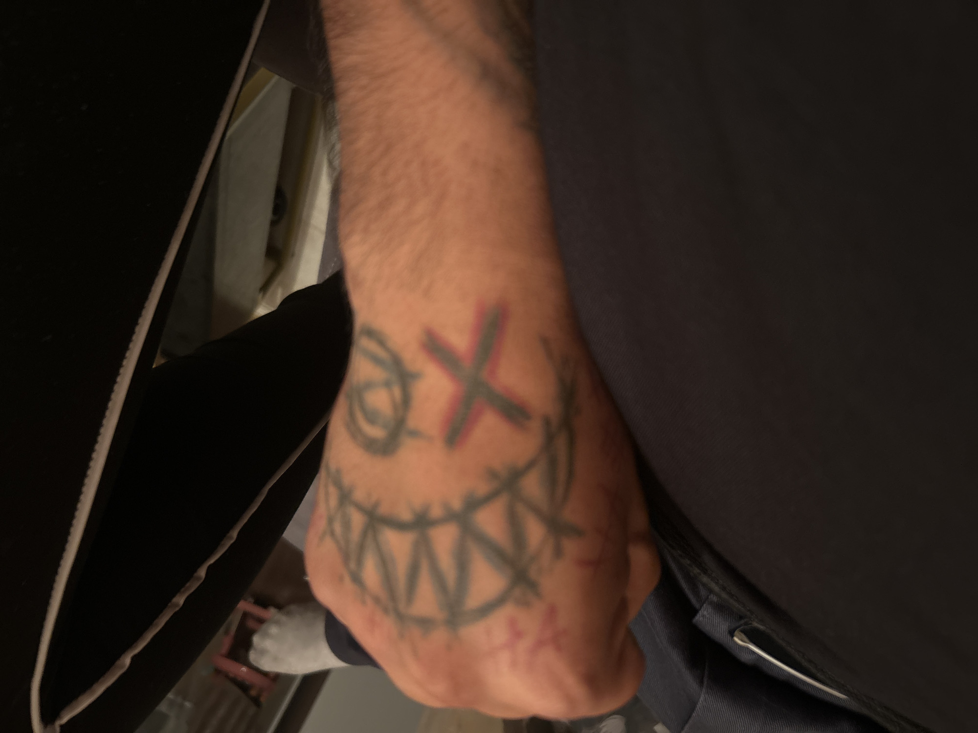 A close-up shot of a right hand resting on a person's leg. The hand has a tattoo of a stylized face with an "X" for eyes and sharp, triangular teeth. The "X" has a reddish hue. The person's leg is covered in a dark-colored fabric, likely clothing. A safety pin is partially visible on the leg garment. A sliver of what appears to be a socked foot can be seen in the lower-left background. The surrounding environment is indistinct. There is no indication of the location being Razhevo Konare, Bulgaria.
