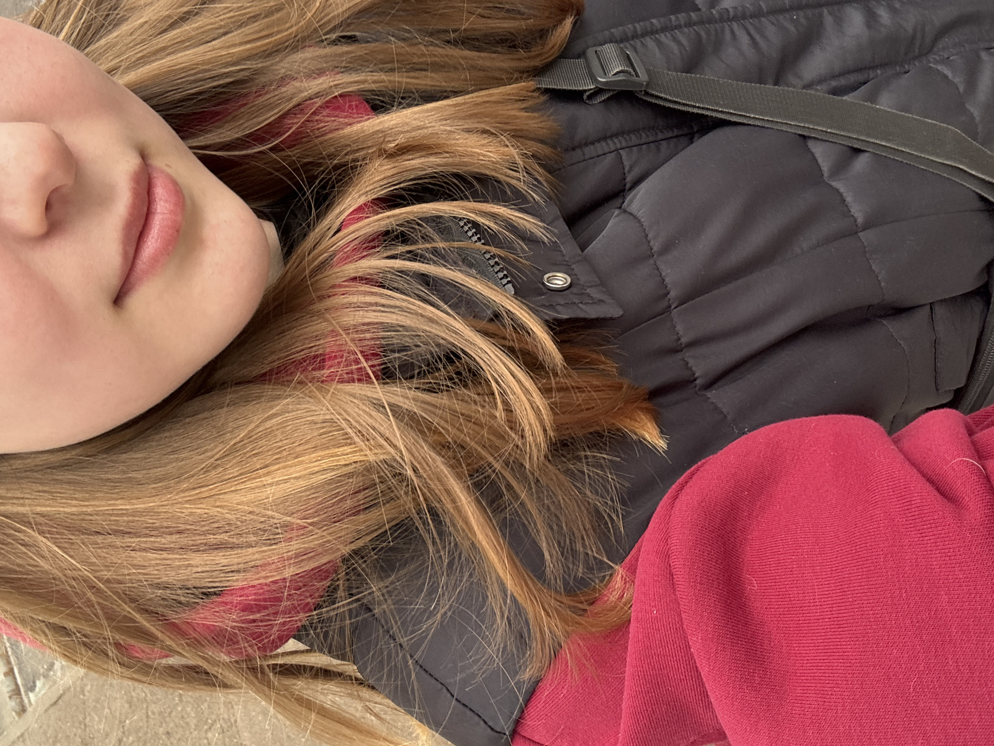 A close-up, angled shot of a person's face with light skin, specifically focusing on the nose and lips. The person is lying down, with their blonde hair spread around their head. The hair appears to be partially covering a black puffer vest, which is partially unzipped. A portion of a red garment, likely a sweatshirt, is also visible. The setting or location of the image cannot be determined solely from this image. There is insufficient data to claim the location to be Chaikovsky, Russia.