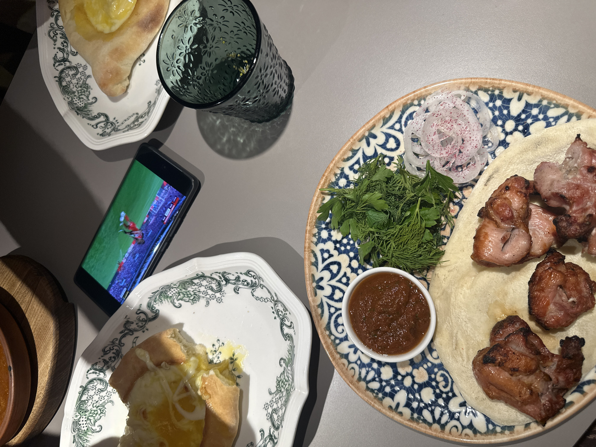 A top-down view depicts a table setting with various items. Two plates, decorated with green floral patterns, hold what appears to be partially consumed khachapuri, a traditional Georgian cheese bread. A smartphone rests near one of the plates displaying a soccer game. Beside the phone is a textured green glass. On the right side is a plate with a blue and white pattern containing shish kebab, herbs (dill), sliced onions with spices, and a small bowl of sauce. The setting suggests a meal or snack is being enjoyed while watching soccer. The location is unknown.