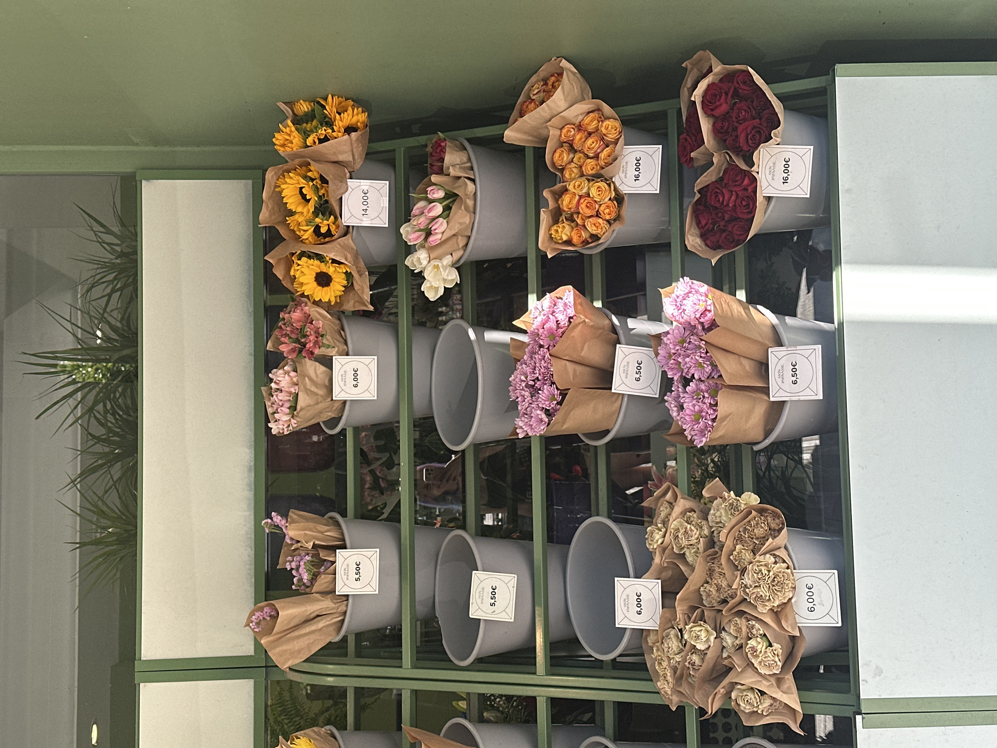 The image shows a flower stand in what appears to be Madrid, Spain. The stand features metal shelving painted green and filled with gray buckets containing various types of flowers wrapped in brown paper. Each bucket is marked with a small price tag. Starting from the top row, there are bouquets of sunflowers, tulips, orange roses, and red roses. The second row includes pink and red flowers, more pink flowers, and another set of pink flowers. The bottom row displays smaller purple flowers, empty gray buckets, tan flowers, and a set of empty gray buckets. The left side of the image is partially obscured by an architectural element and some greenery. The pricing tags on the buckets display prices like 5.50€, 6.00€, 6.50€, 14.00€, and 16.00€. Some tags include the word "PARNASSE."