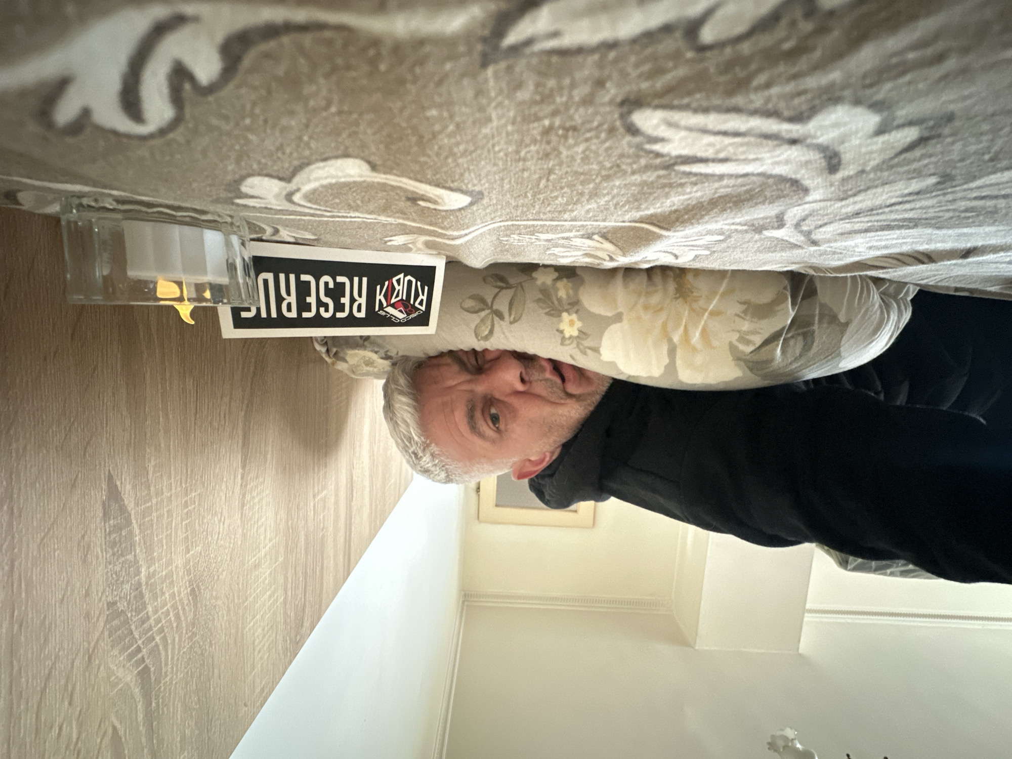 A disoriented view shows a man's head wedged between a floral pillow and a patterned surface, presumably a bed or sofa. The man has gray hair and appears to be wearing a dark jacket. A "Ruby Disco Club Reserve" sign and a lit candle in a glass holder are positioned near his head. The background includes a light-colored wall and wooden flooring. There is no indication of Burgas, Bulgaria in the image.