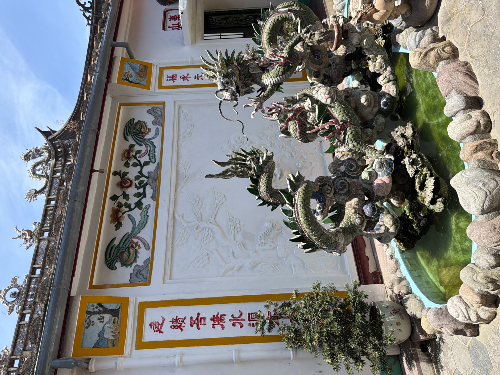 In Hội An Ward, Vietnam, a vibrant courtyard features a decorative fountain with two intricately designed ceramic dragons. The dragons, predominantly green with colorful accents, are perched atop a rocky structure within a turquoise pool. The pool is surrounded by a border of rough, grey and pink stones. To the left of the fountain, a small, potted evergreen tree stands next to a white wall adorned with red Chinese calligraphy. Above the wall, a rooftop with ornate dragon motifs and tiled eaves is visible against a clear blue sky. The wall itself is decorated with bas-relief sculptures of birds and flowers, as well as framed paintings depicting a white animal in a pastoral scene and a mythical bird.