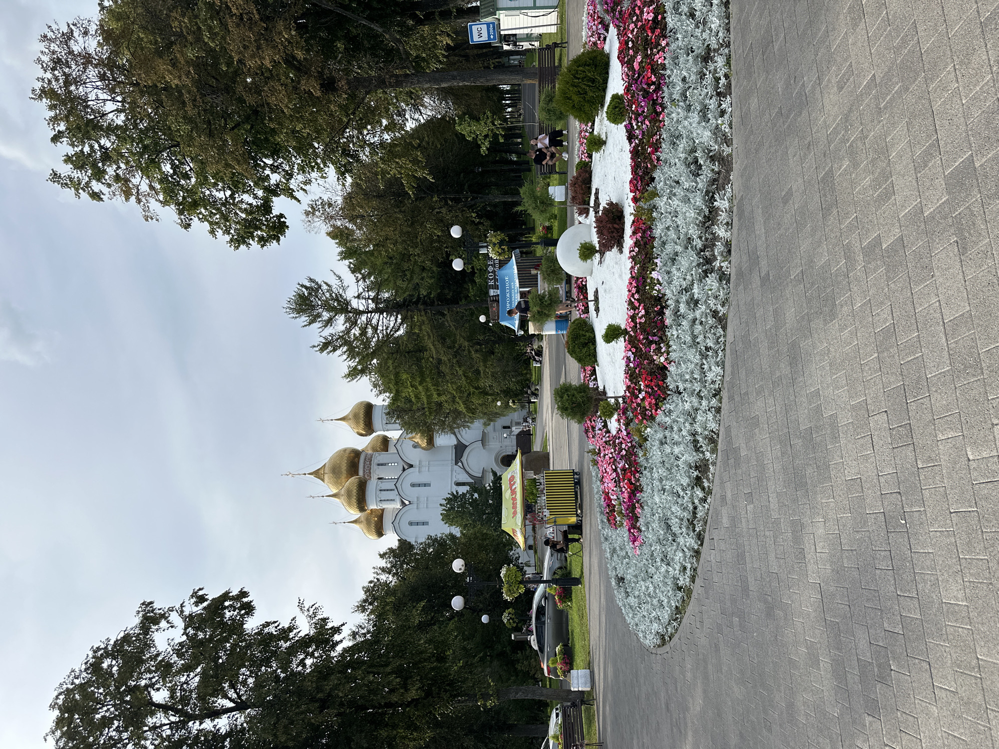 A wide-angle, eye-level shot captures a public park scene in Rusanovo, Russia, on an overcast day. In the background, a large white Orthodox cathedral with multiple golden domes stands prominently. The foreground features a curved, paved walkway made of gray interlocking bricks, leading towards a circular flower bed. This bed is densely planted with a variety of colorful flowers, primarily pink and red, bordered by silver-leafed plants. Beyond the flower bed, a decorative white mound with small evergreen bushes and a few bare branches is visible. To the left of the flower bed, a yellow and black striped kiosk sells ice cream, with a person sitting on a stool beside it. Further back, cars are parked on the grass, and several people are seated on benches or walking along the park's paths, surrounded by lush green trees. A sign for a "WC" (water closet) is visible on the right side of the image, mounted on a white structure. The overall atmosphere is serene and suggests a pleasant, well-maintained public space.