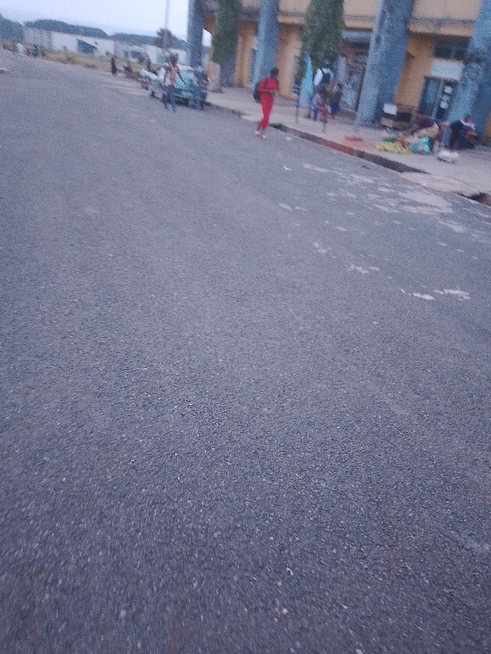 The image shows a street scene in Ogboko, Nigeria. The foreground is dominated by an asphalt road, while in the background there are buildings with large pillars and a few people. There is a person wearing a red jumpsuit walking along the sidewalk. Further back, there are several individuals gathered near the building, some appearing to be sitting or standing. A vehicle is also visible nearby. Some items that could be merchandise are visible near the building.