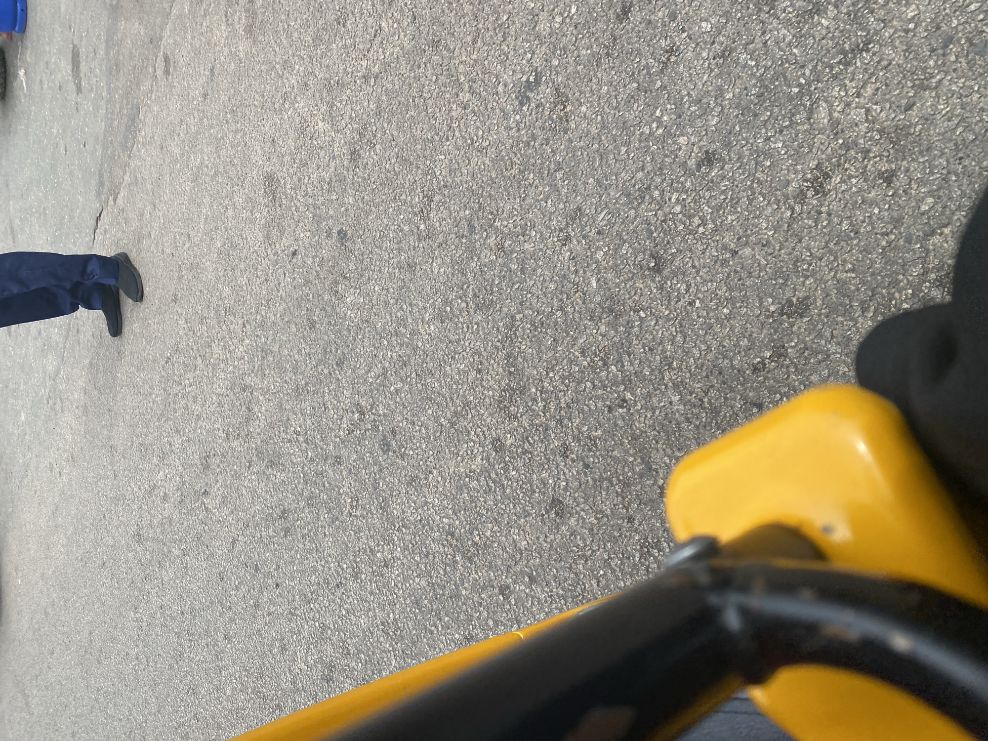 The image shows a perspective from a yellow and black object, possibly a vehicle or a piece of equipment, looking out onto an asphalt surface. A person's legs, clad in dark blue trousers and black shoes, are visible in the upper right portion of the frame. The asphalt appears worn and textured. The scene is likely outdoors, given the lighting and the texture of the ground. The specific location in Jalingo, Nigeria, cannot be determined from the visual information provided.