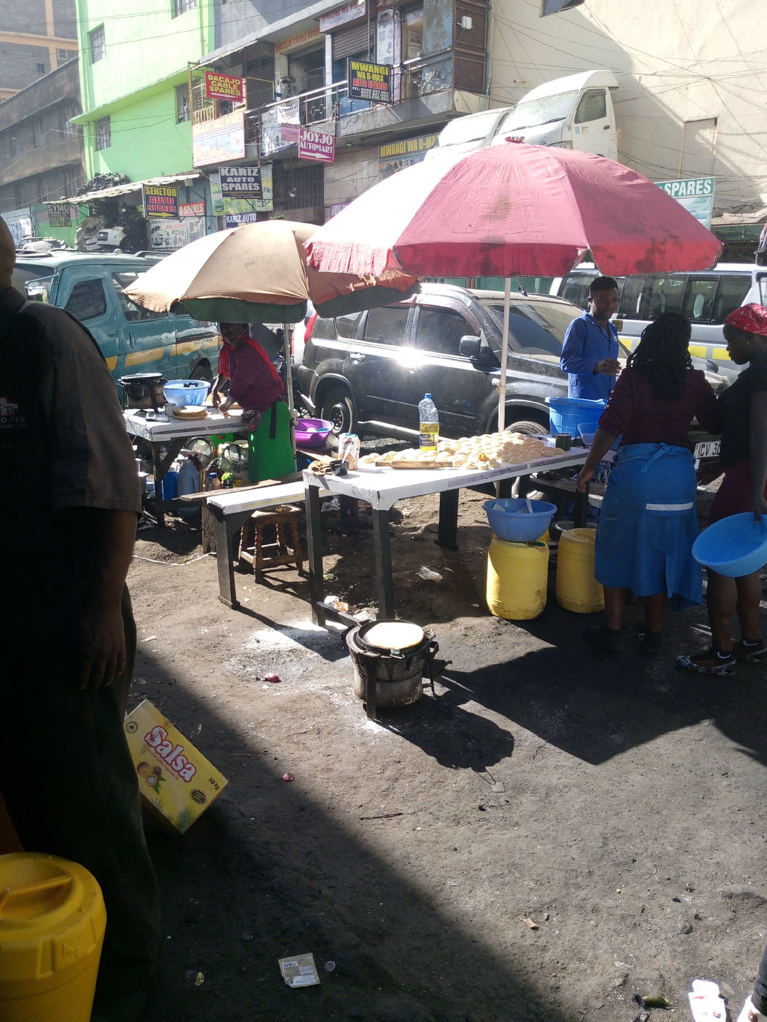 Here's a detailed description of the image:
**Scene:** An outdoor food vending setup on a street in Nairobi, Kenya. The ground is dirt and asphalt with trash scattered around.
**People:** Several individuals are present. One person stands partially in the foreground to the left, obscuring the view. There are at least four women actively involved in the food preparation and service. One man stands nearby.
**Objects:**
* Two tables are set up as workstations, covered by large sun umbrellas (one tan, one red).
* Food items, likely chapati or similar flatbread, are displayed on the tables.
* There are yellow plastic containers (likely for water or ingredients).
* Blue plastic bowls are present.
* A small charcoal stove or burner is on the ground with a pot on top.
* A box of "Salsa" (cooking oil) is visible on the ground.
* Various vehicles are parked in the background, including cars and vans.
**Actions/Interactions:**
* The women are actively preparing and serving food.
* The man stands by in a blue shirt.
* Money is exchanged between the customers and the vendors.
**Environment:**
* The scene is urban, likely a commercial area.
* Buildings with signs (auto parts stores) are visible in the background. The OCR reads: "DAKAJO CABLE SPARES", "KARIZ AUTO SPARES", "JOJO AUTOMART", "SENATOR AUTO SPARES".
* The weather appears sunny, with harsh shadows.